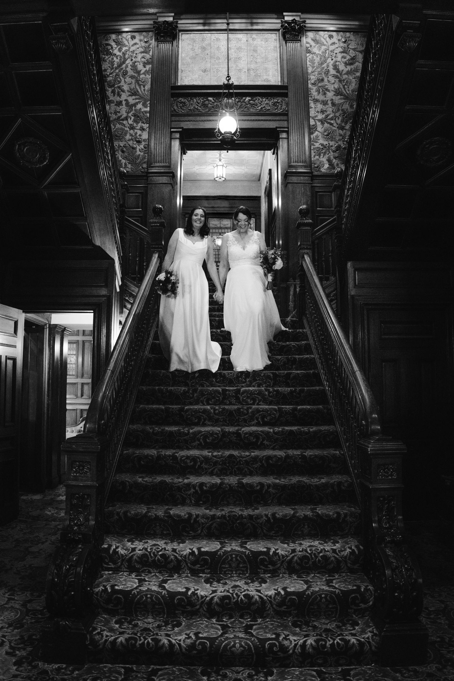 Two brides in white gowns descend an ornate wooden staircase in a grand historic venue, black and white photo.