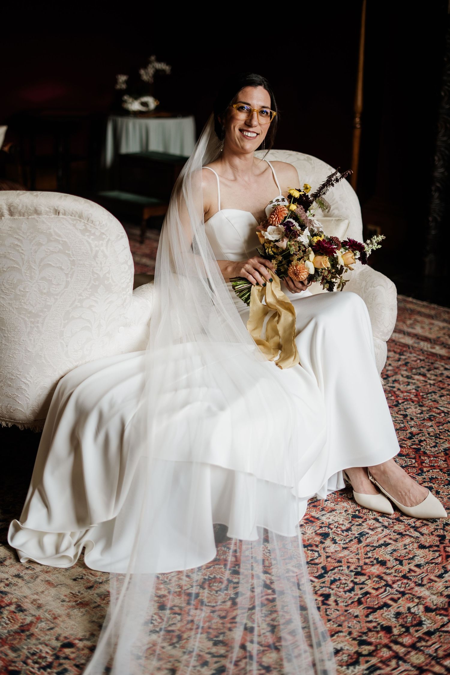 Smiling bride in white gown and veil sits on vintage chair holding colorful bouquet on patterned rug.