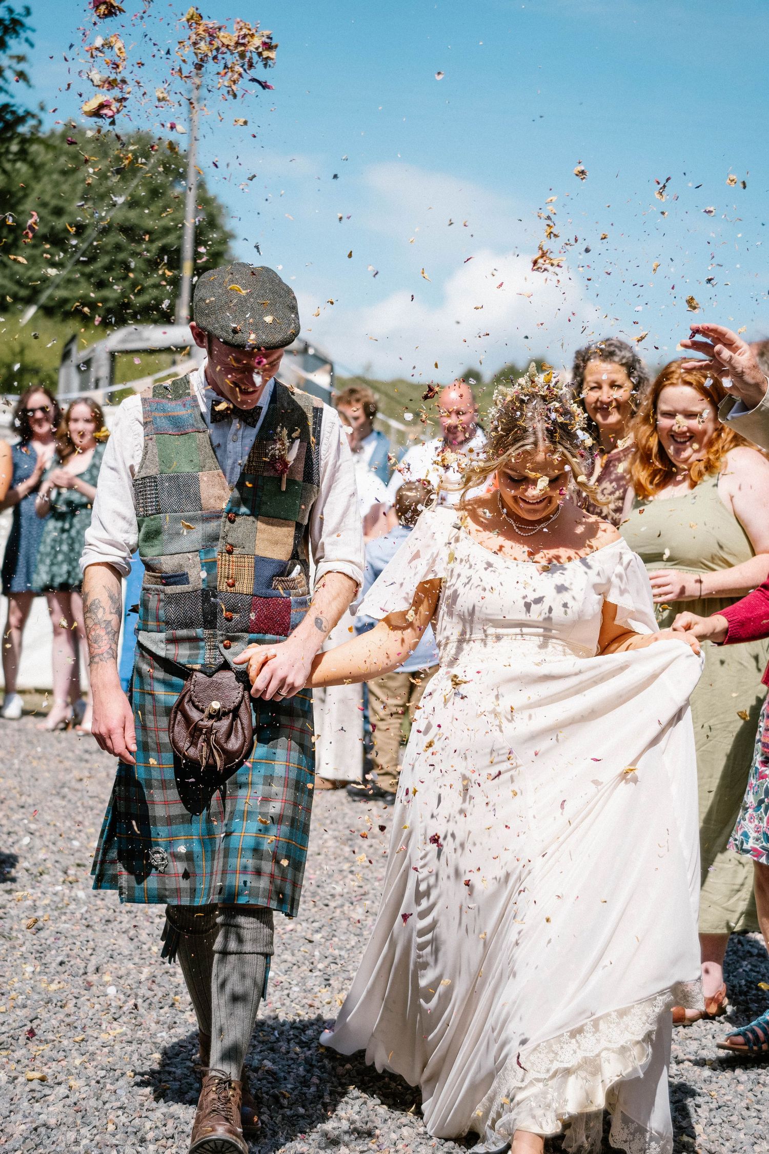 Wedding guests shower the newlyweds with confetti as they walk down a path in Scottish highland dress.