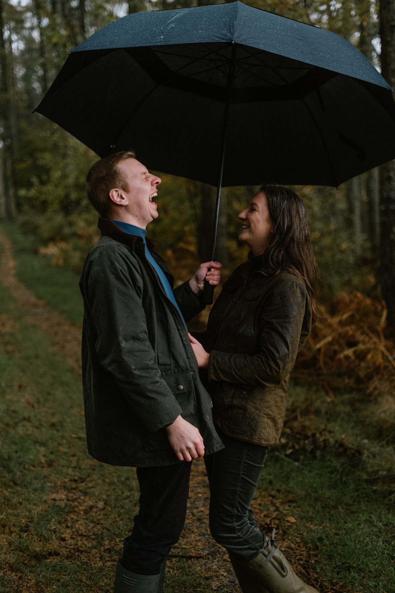 A playful moment shared under black umbrellas in a wooded autumn setting.