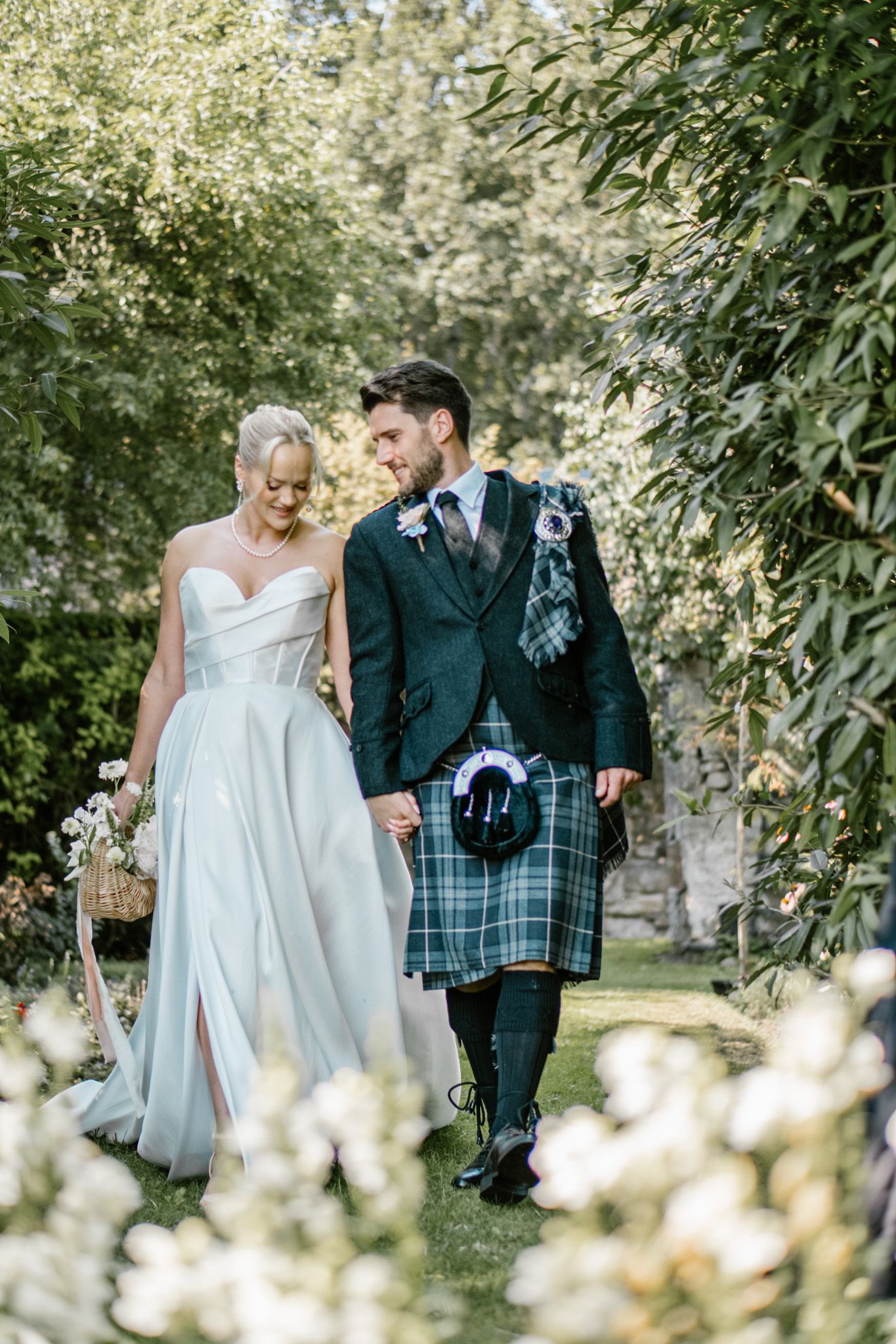 Couple walking through garden path with white flowers, groom wearing traditional Scottish kilt.