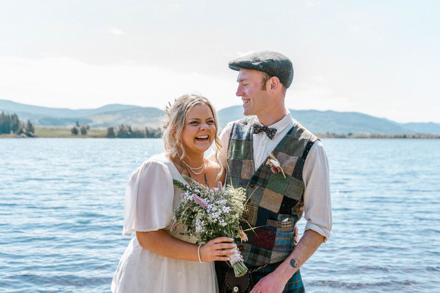 Couple enjoying champagne by the water in Scottish wedding attire.