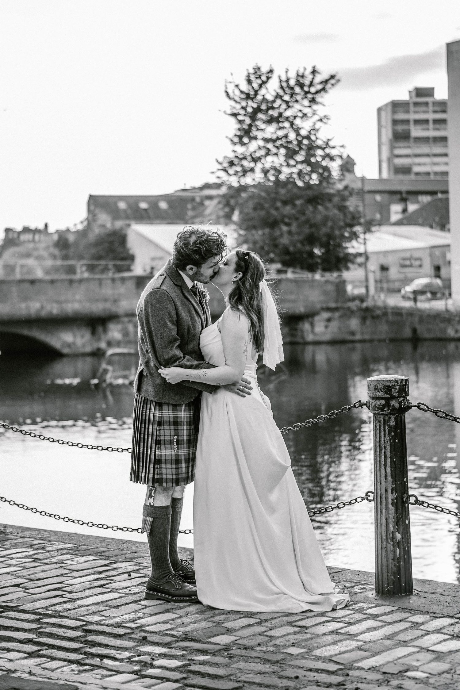 A romantic black and white wedding photo by a canal with traditional Scottish attire.