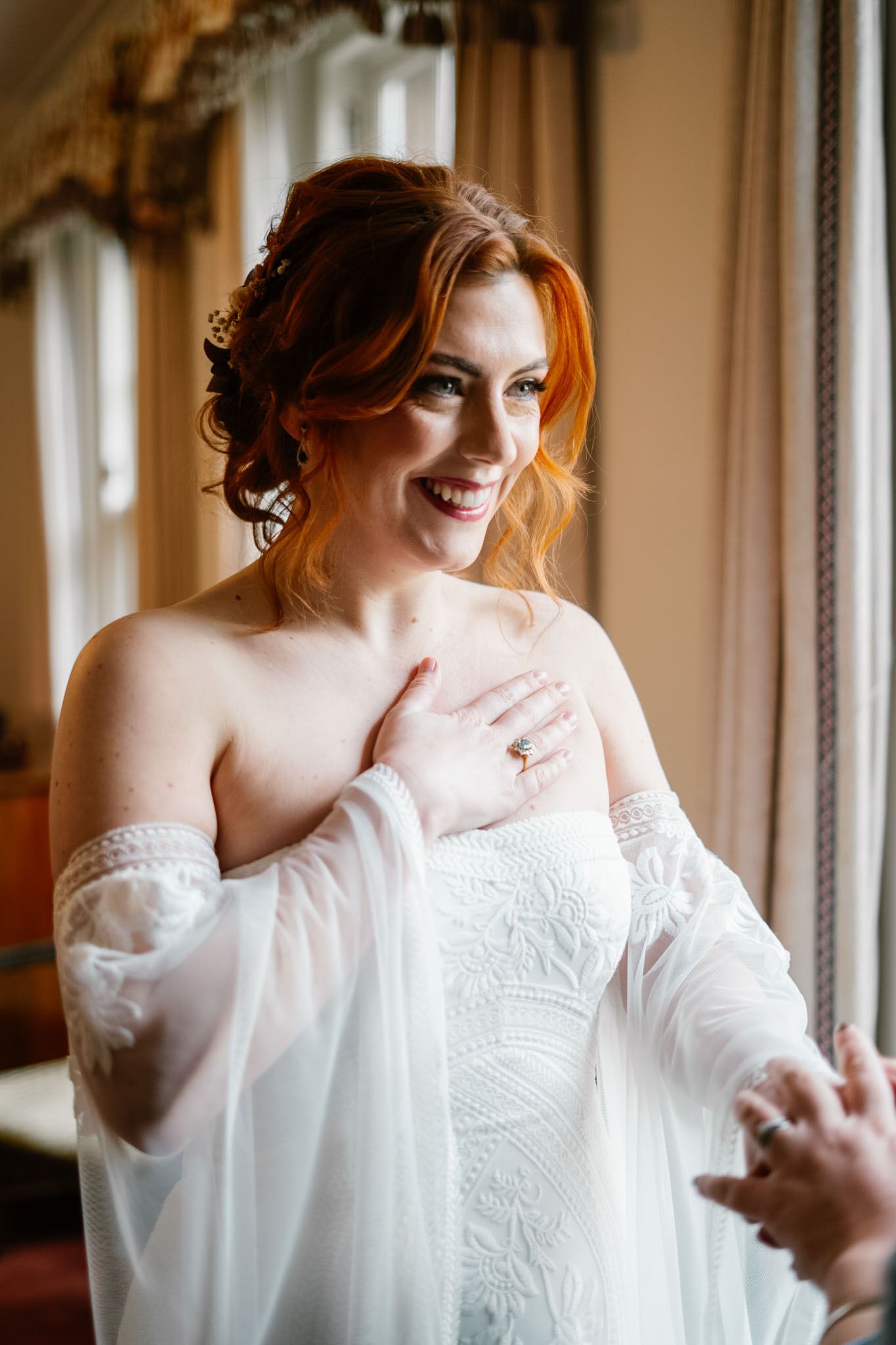 Woman in white lace robe preparing for wedding day by window.