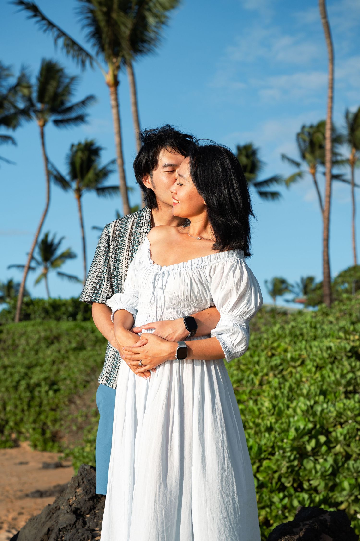 Couple in a tender embrace on a tropical beach, the woman in a white dress, with palm trees and a blue sky behind them.