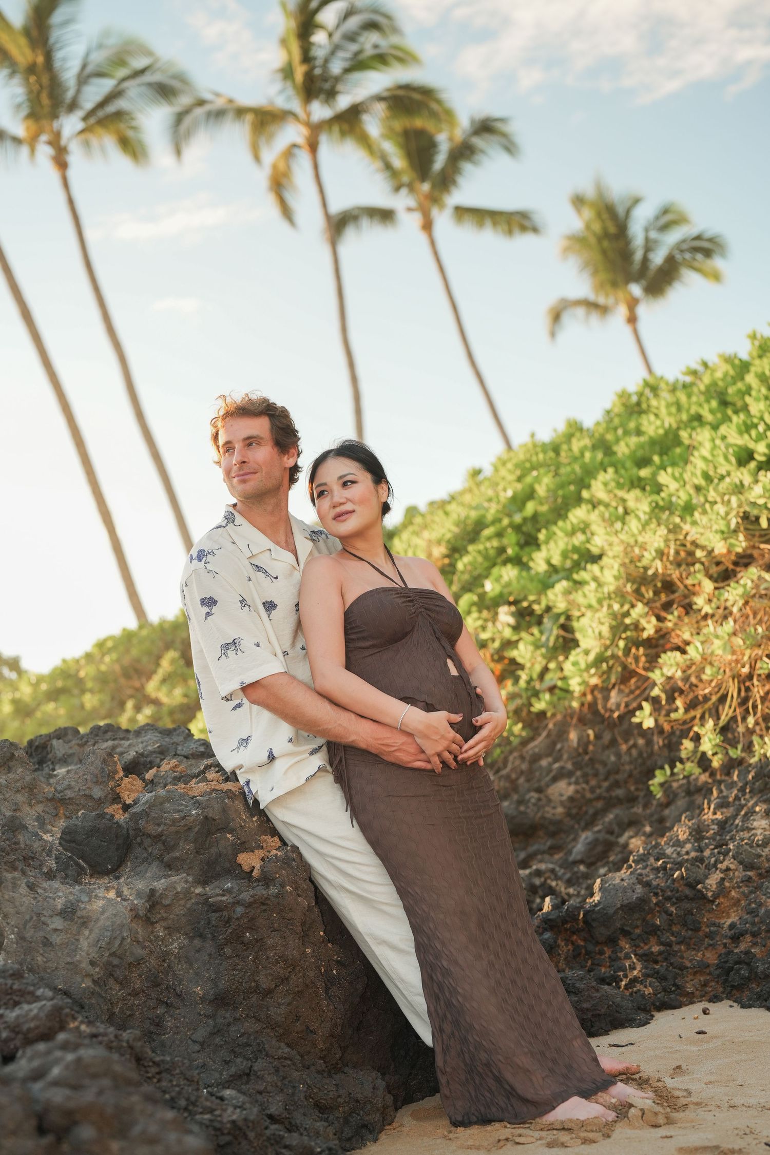Expectant couple embracing on a rocky tropical beach at sunset, with palm trees and green coastal shrubs in the background.