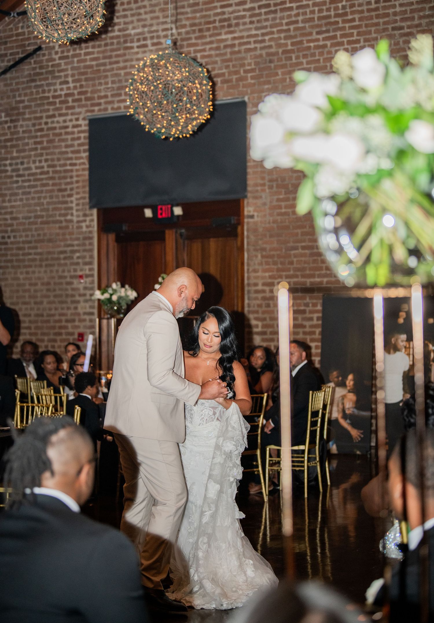 Dancing couple illuminated by hanging lights in a rustic brick-walled reception venue.