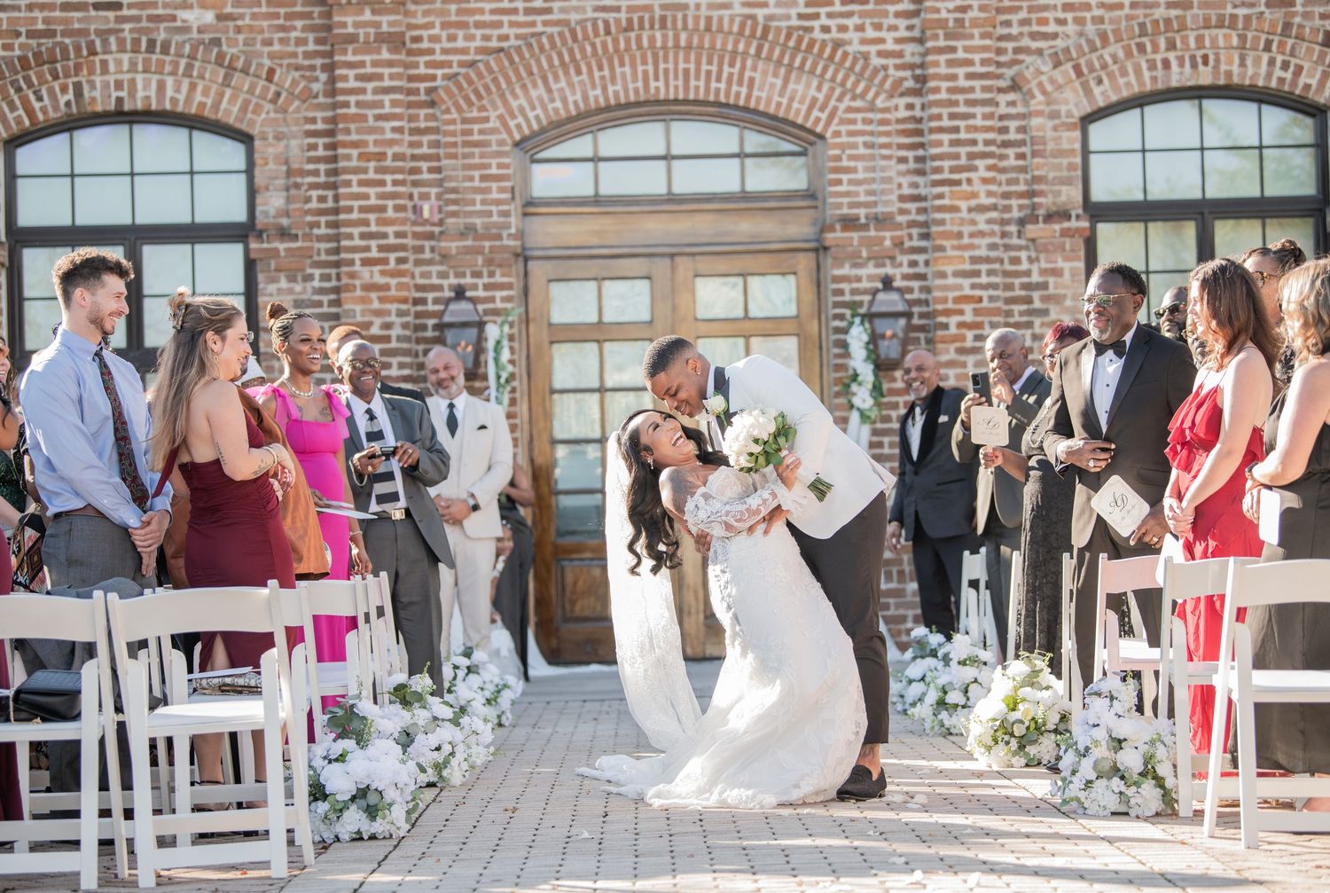 Wedding venue panorama showing guest seating and floral arrangements against brick building.