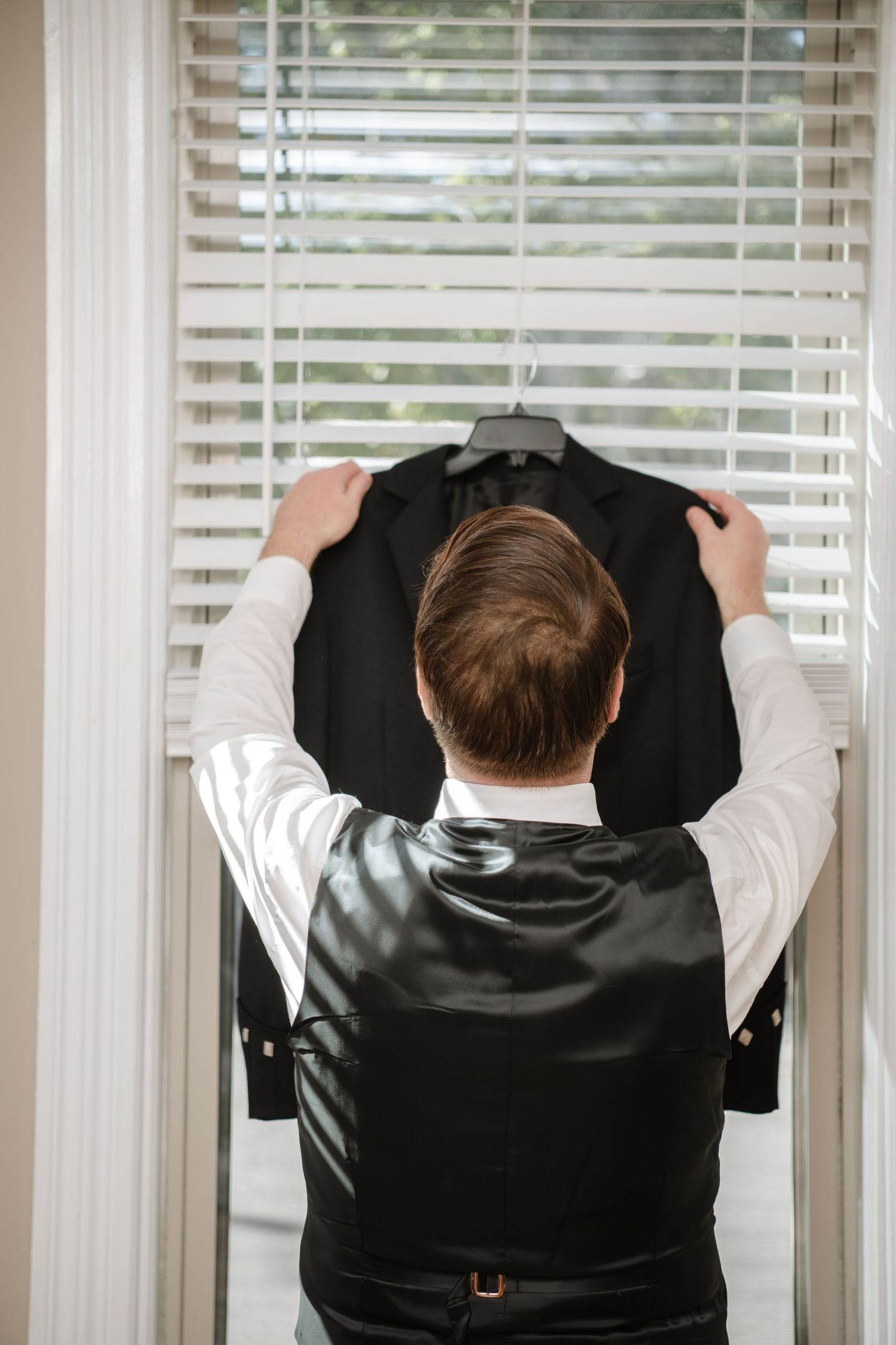 A person in formal attire adjusts a black suit jacket while standing near venetian blinds.