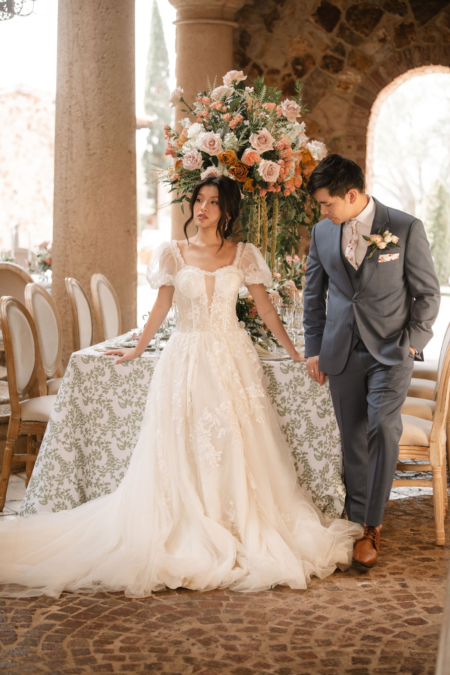 Wedding couple poses elegantly among white chairs in a rustic stone venue.