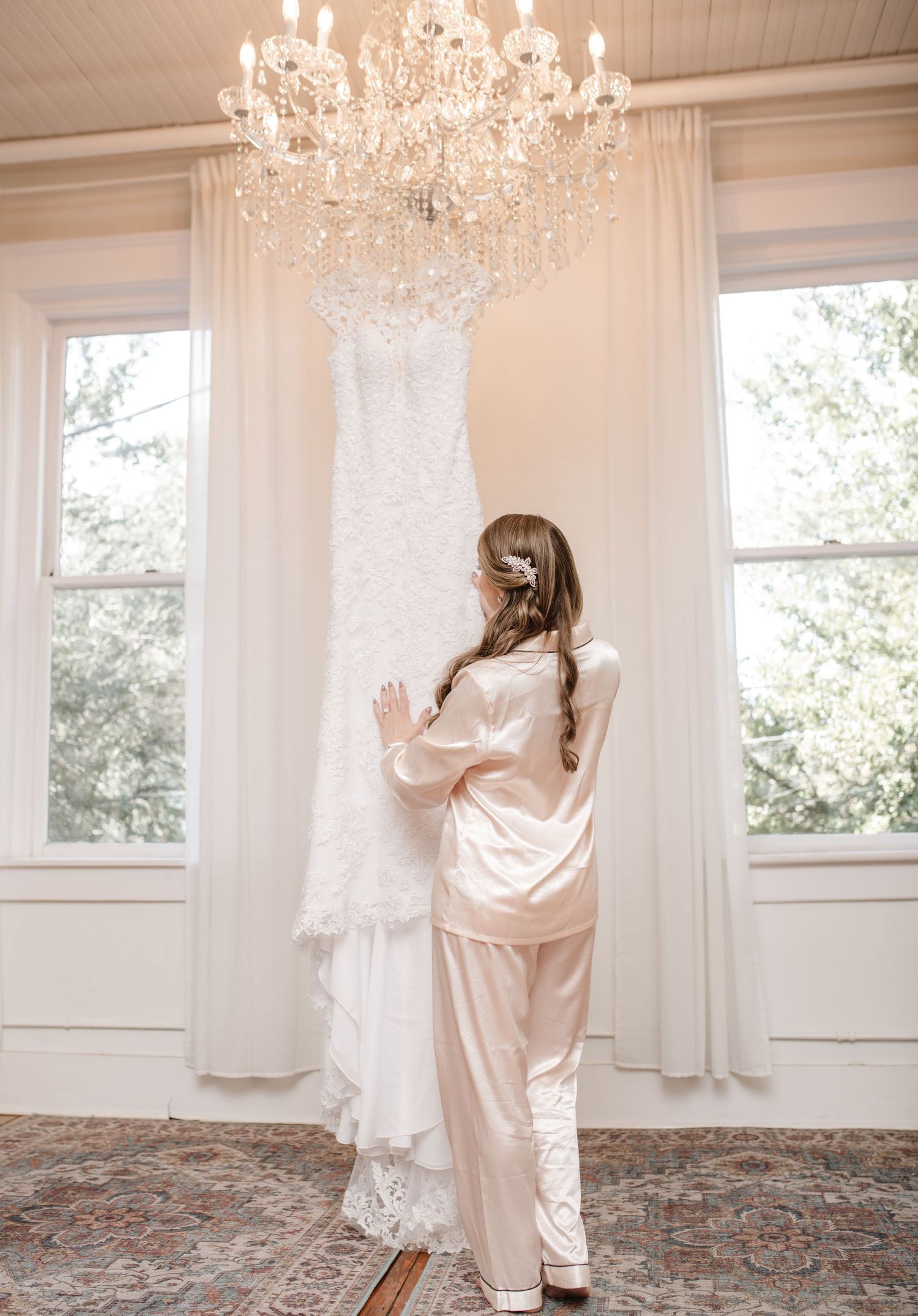 White lace wedding dress hanging from crystal chandelier in bright sunlit room.