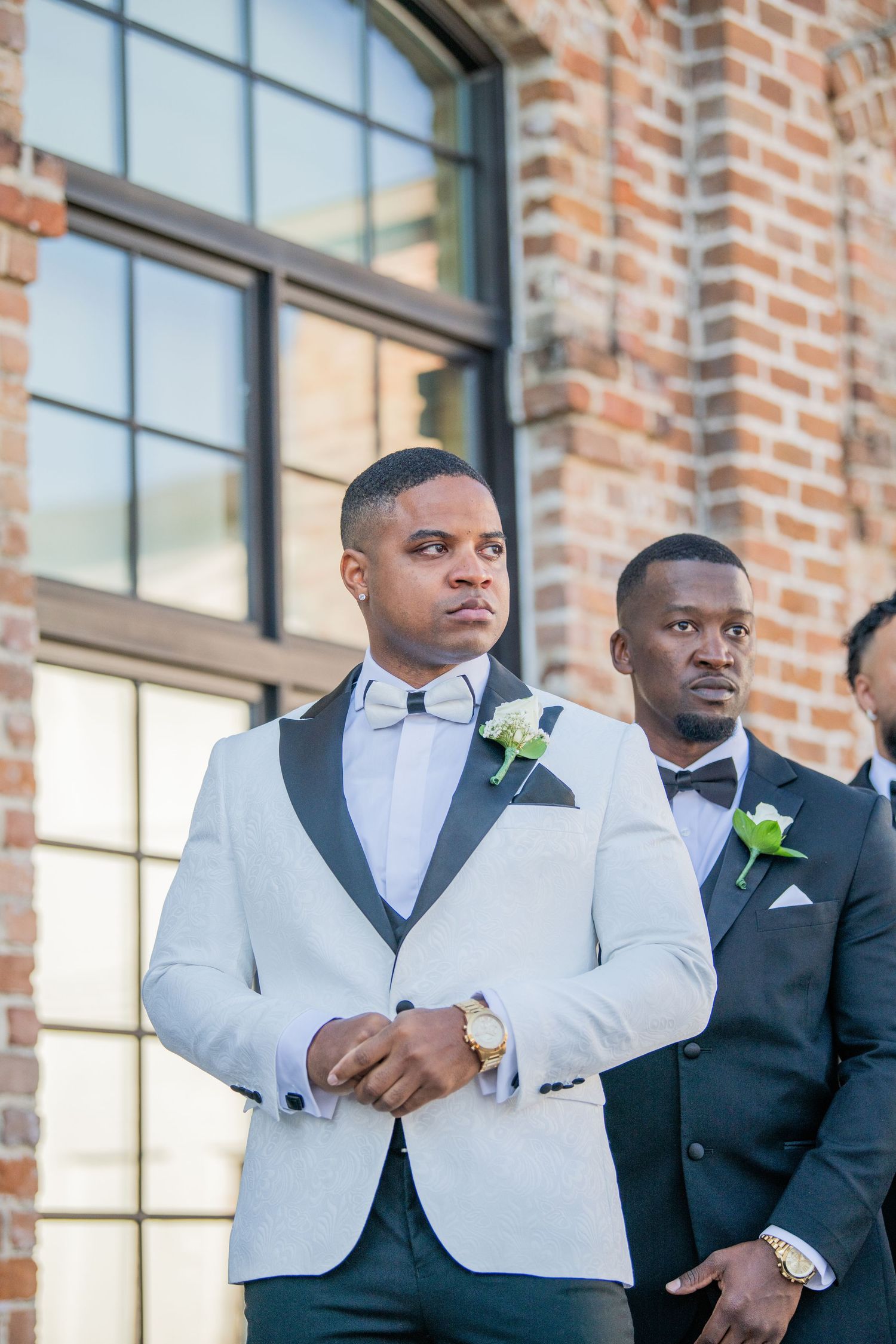 Groomsmen in formal attire pose against a brick wall with large windows.