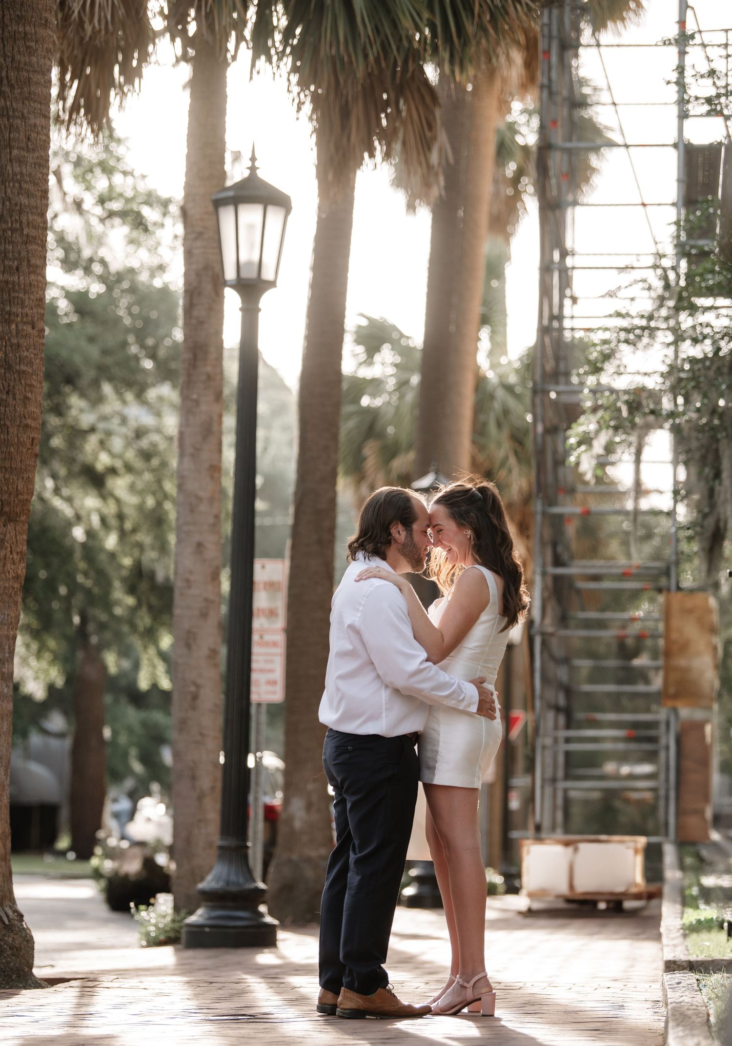 A romantic couple shares an intimate moment among palm trees in a sun-dappled outdoor setting.