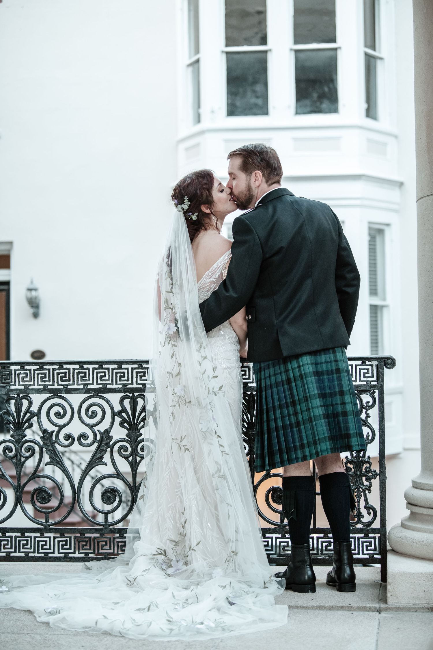 A romantic wedding moment captured on a balcony with ornate railings featuring Greek key patterns.