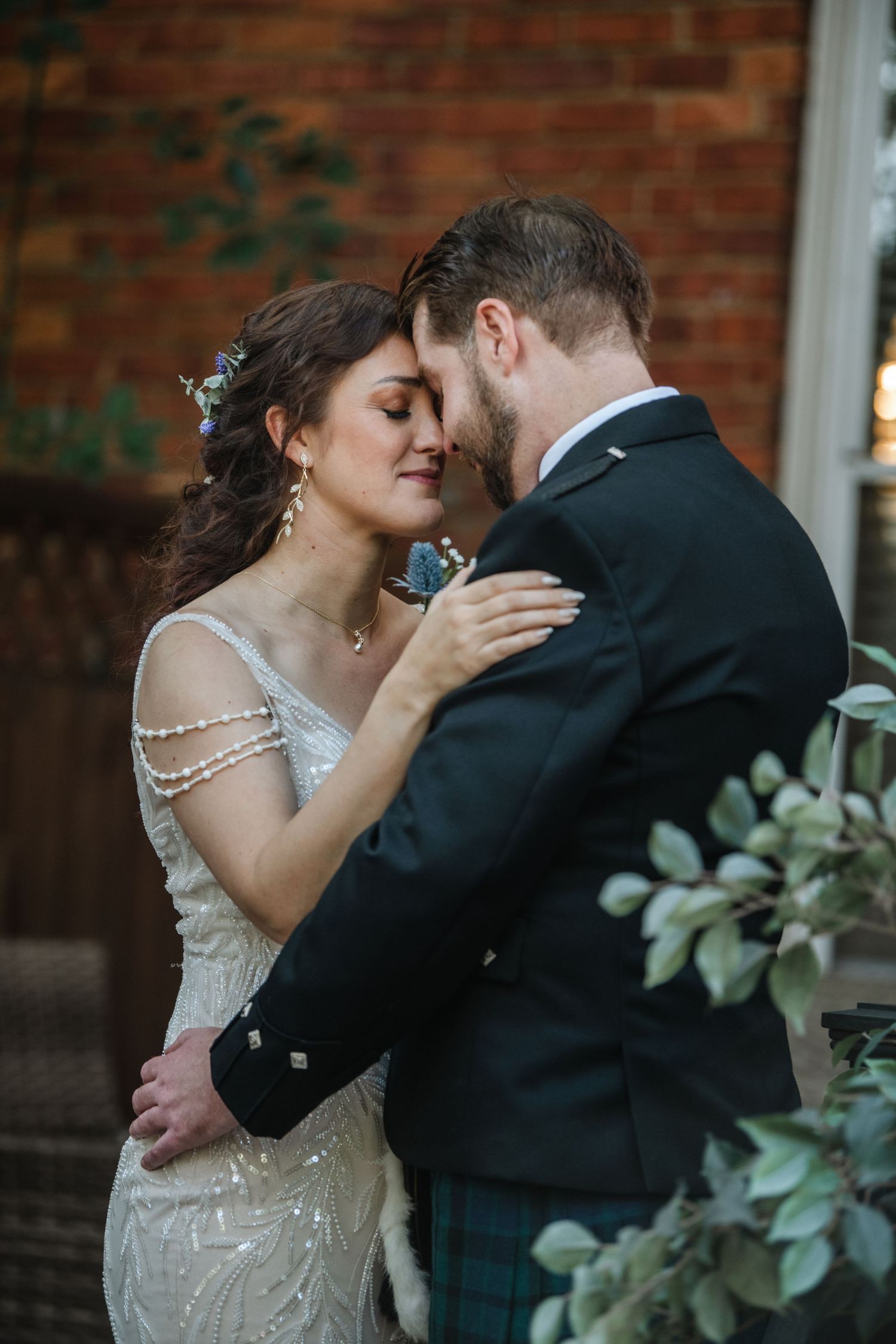 Romantic moment between couple in elegant wedding attire against brick wall with greenery.