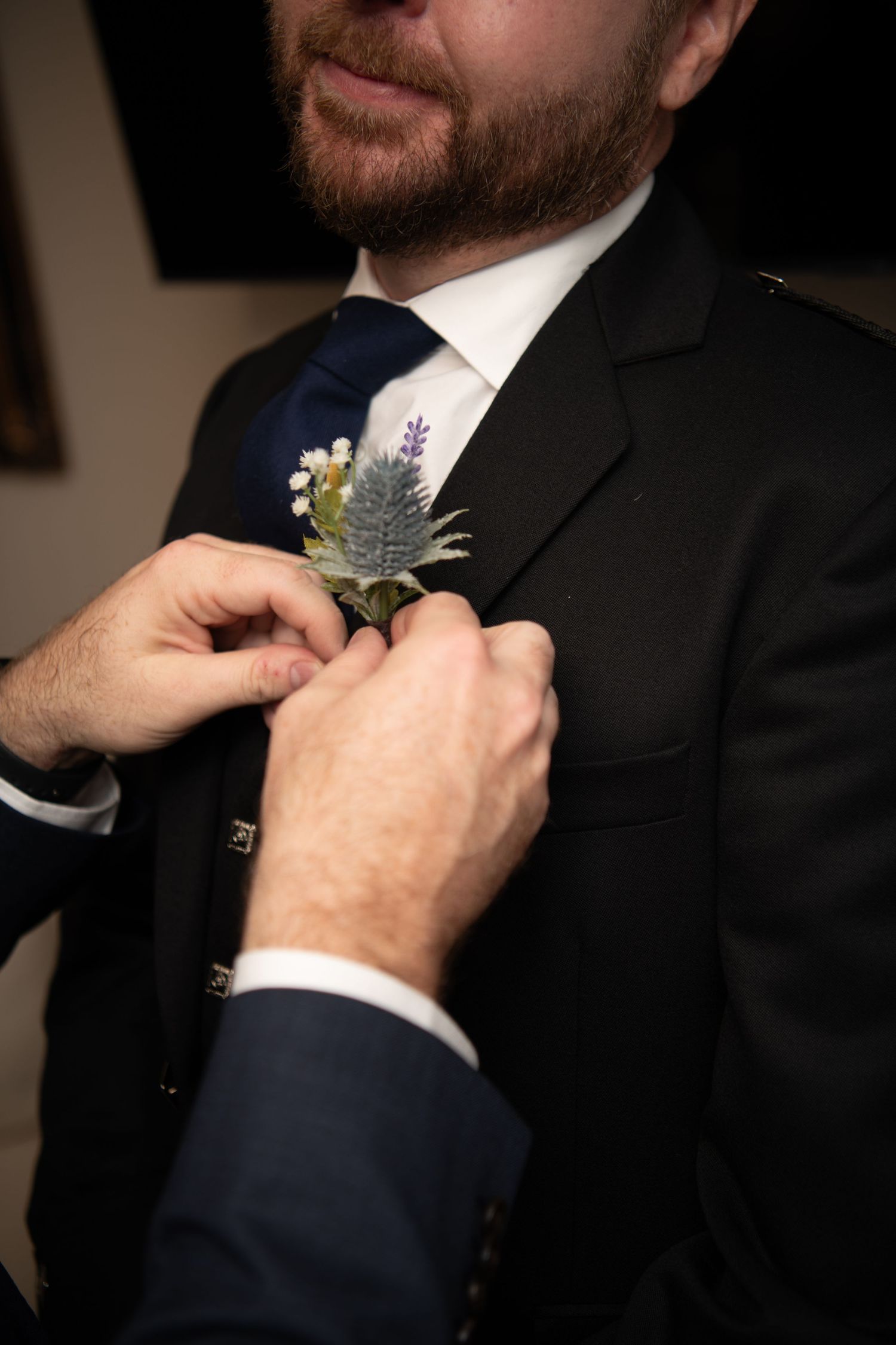 Close-up of hands pinning a boutonniere with thistle and small flowers to a dark suit lapel.