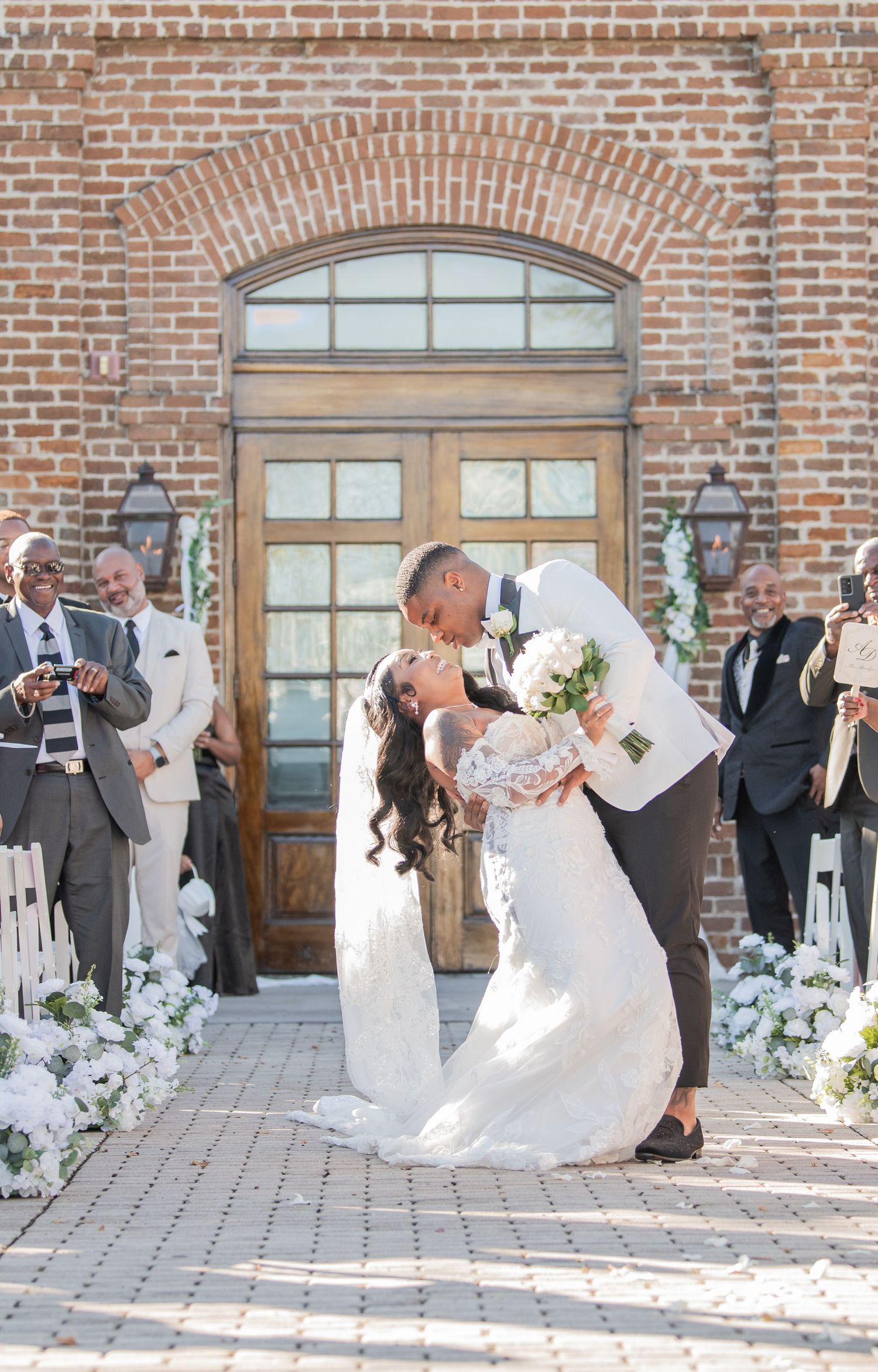 A romantic wedding kiss outside a rustic brick church entrance with white floral decorations.