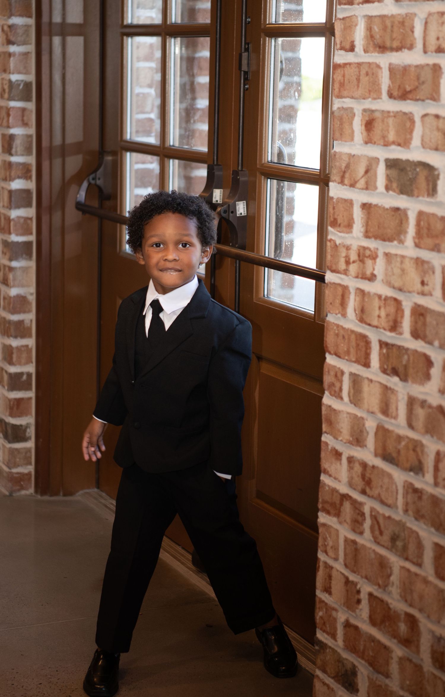 Two small children in formal black suits pose playfully in front of wooden doors.