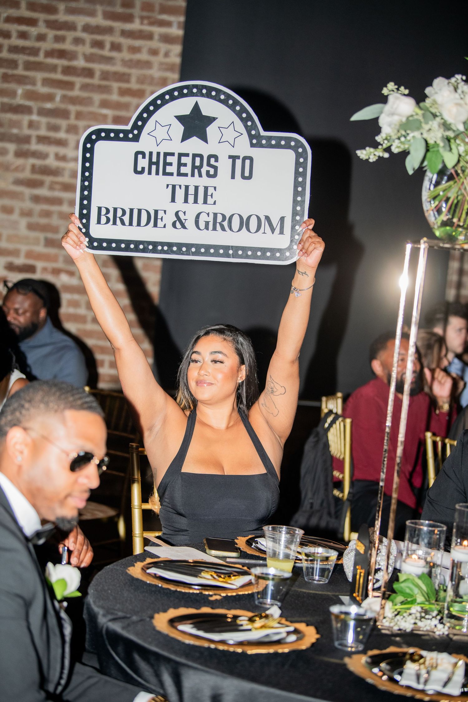 Wedding guest holds up a 'Cheers to the Bride & Groom' sign at reception dinner.