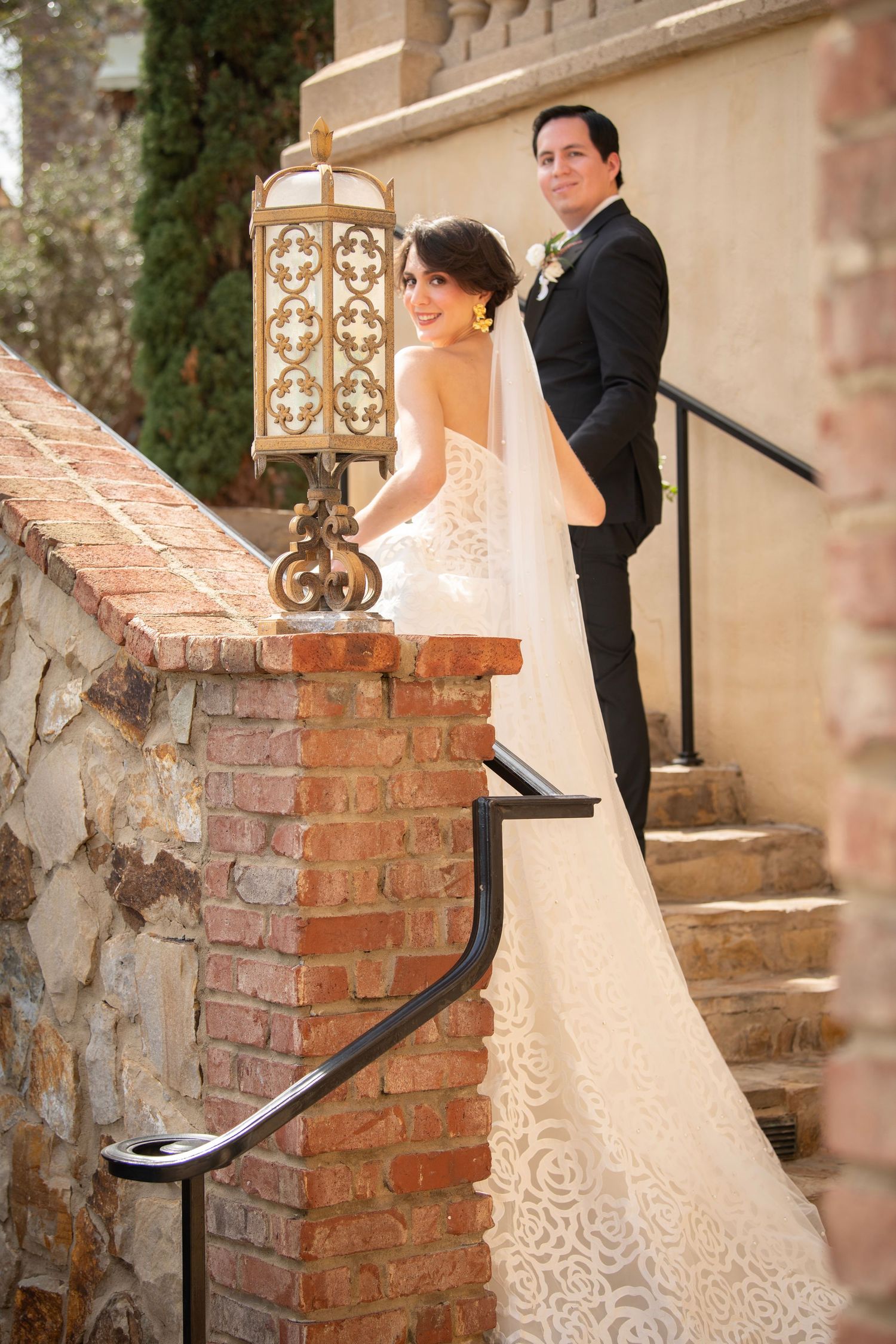 Bride in detailed lace gown posing on vintage brick steps at wedding venue.
