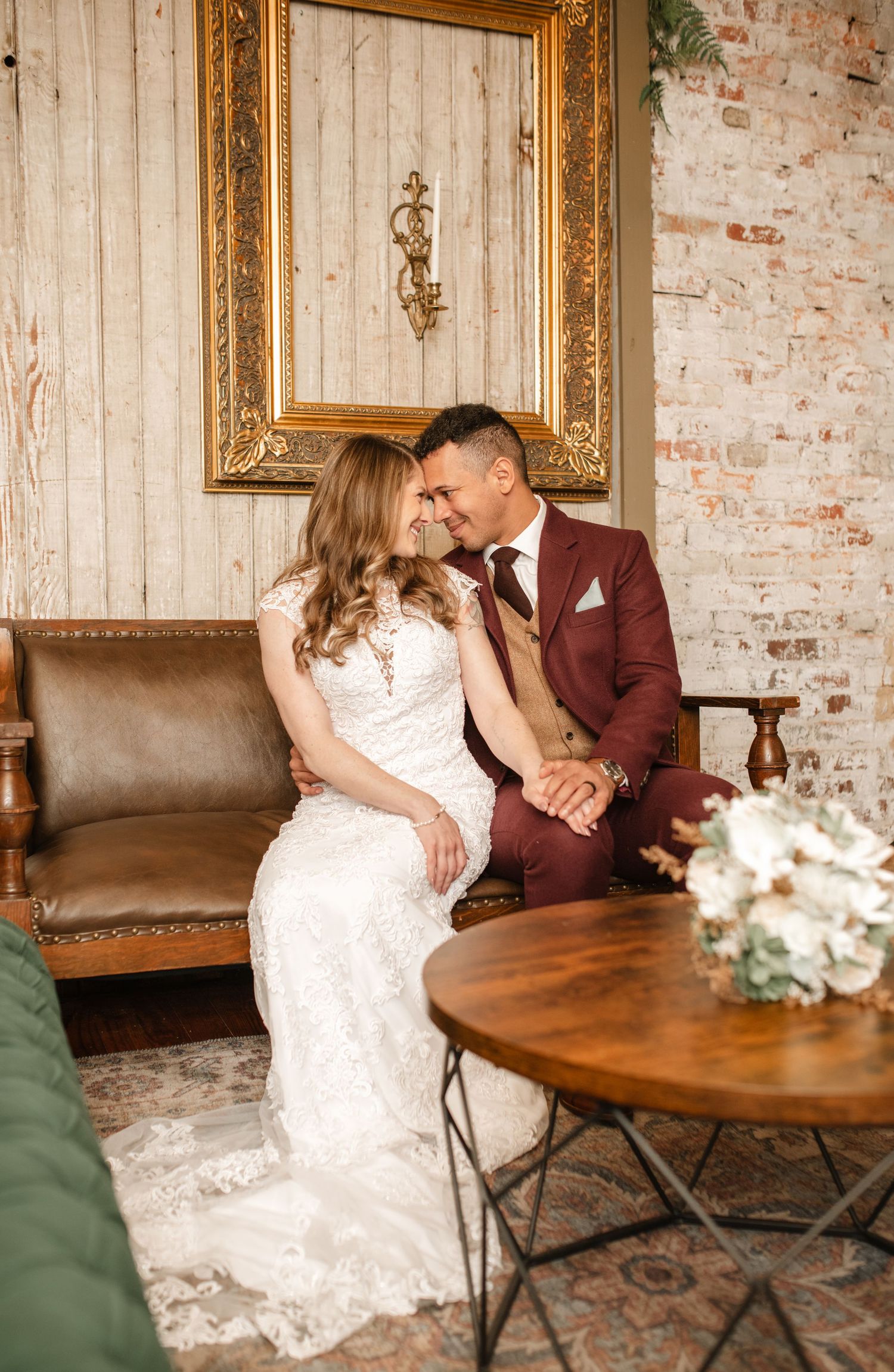 Wedding couple poses intimately on vintage leather furniture against exposed brick walls.