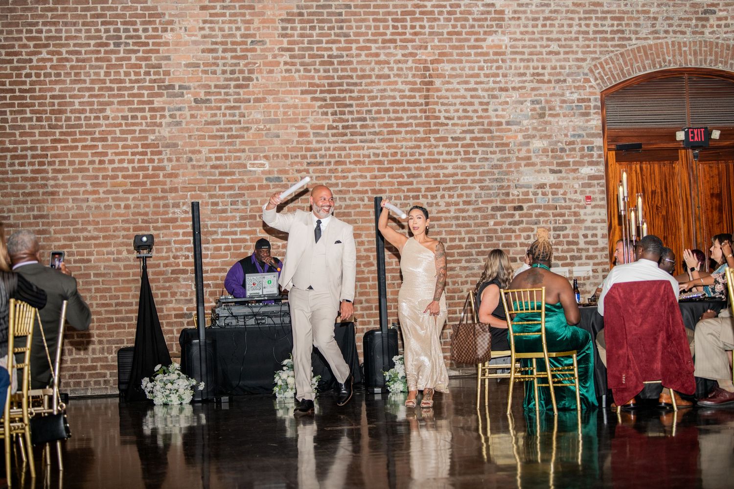 Wedding reception guests dancing and celebrating in a rustic brick venue with gold chairs.