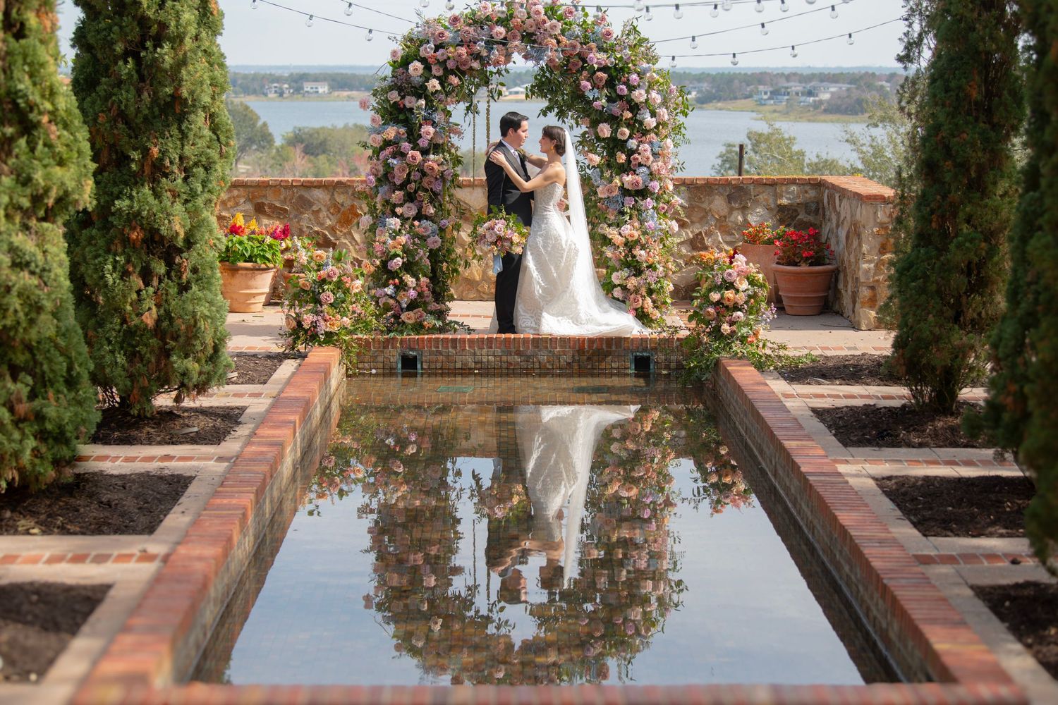 Outdoor wedding venue featuring reflecting pool and floral arch with string lights overhead.