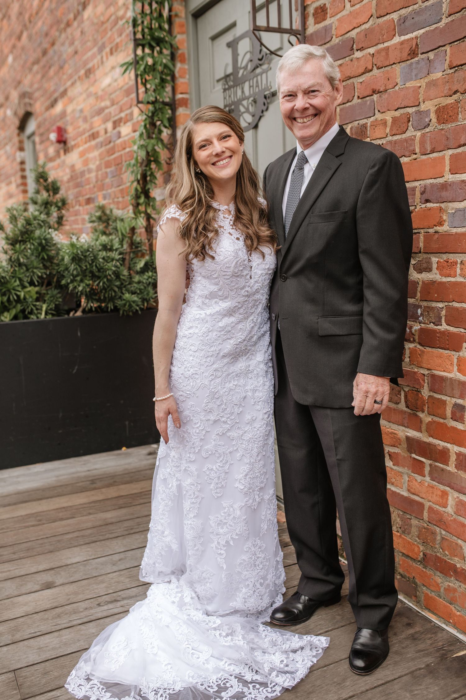Wedding couple poses together against exposed brick wall and greenery.