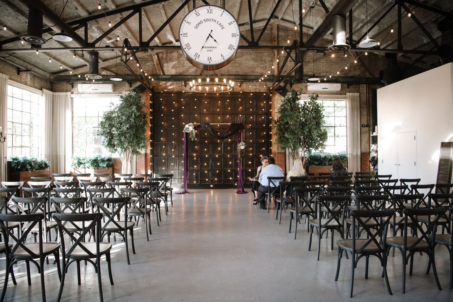 Modern industrial wedding venue with string lights and cross-back chairs set for ceremony.