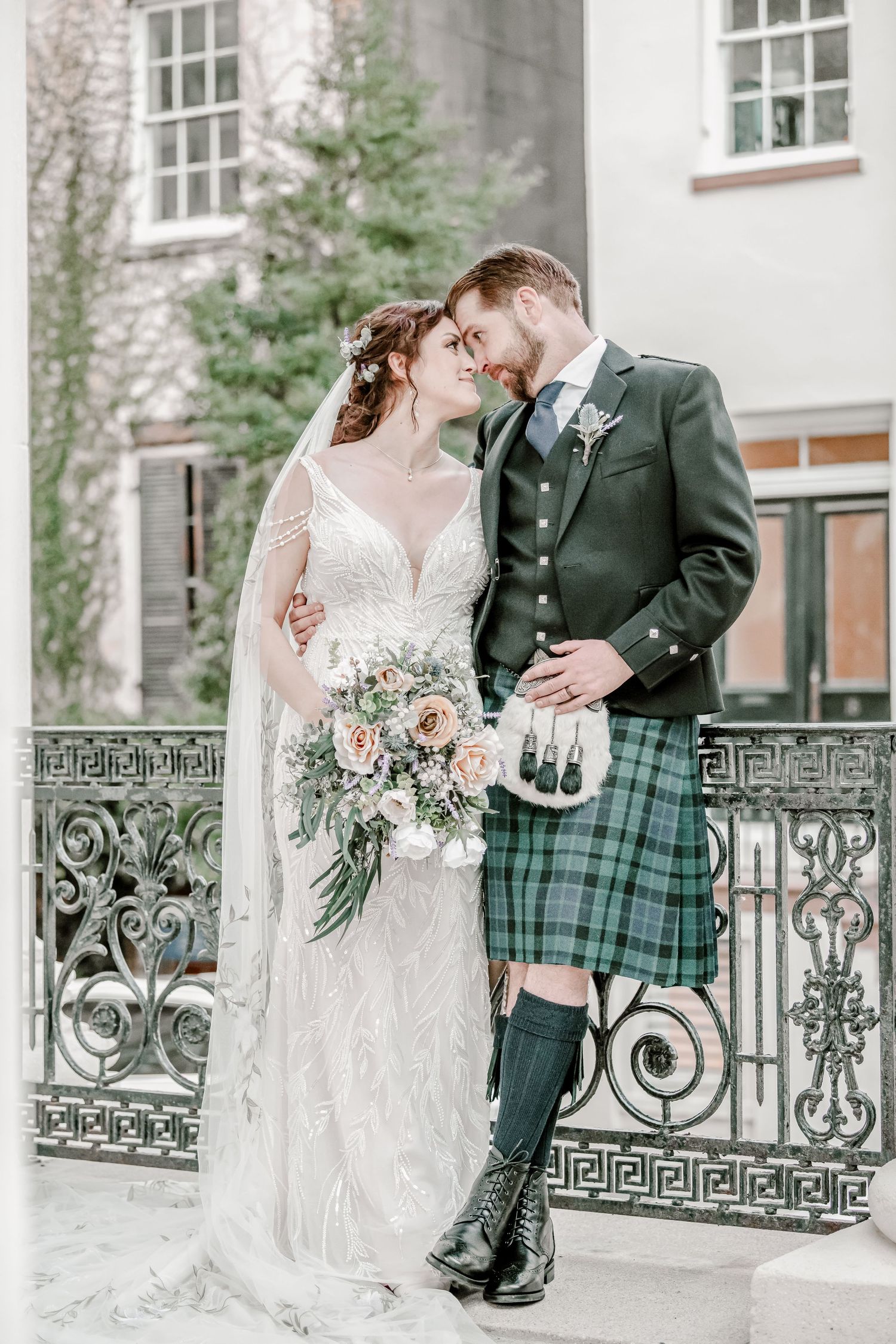 A romantic wedding couple in traditional Scottish attire pose against an ornate iron railing.
