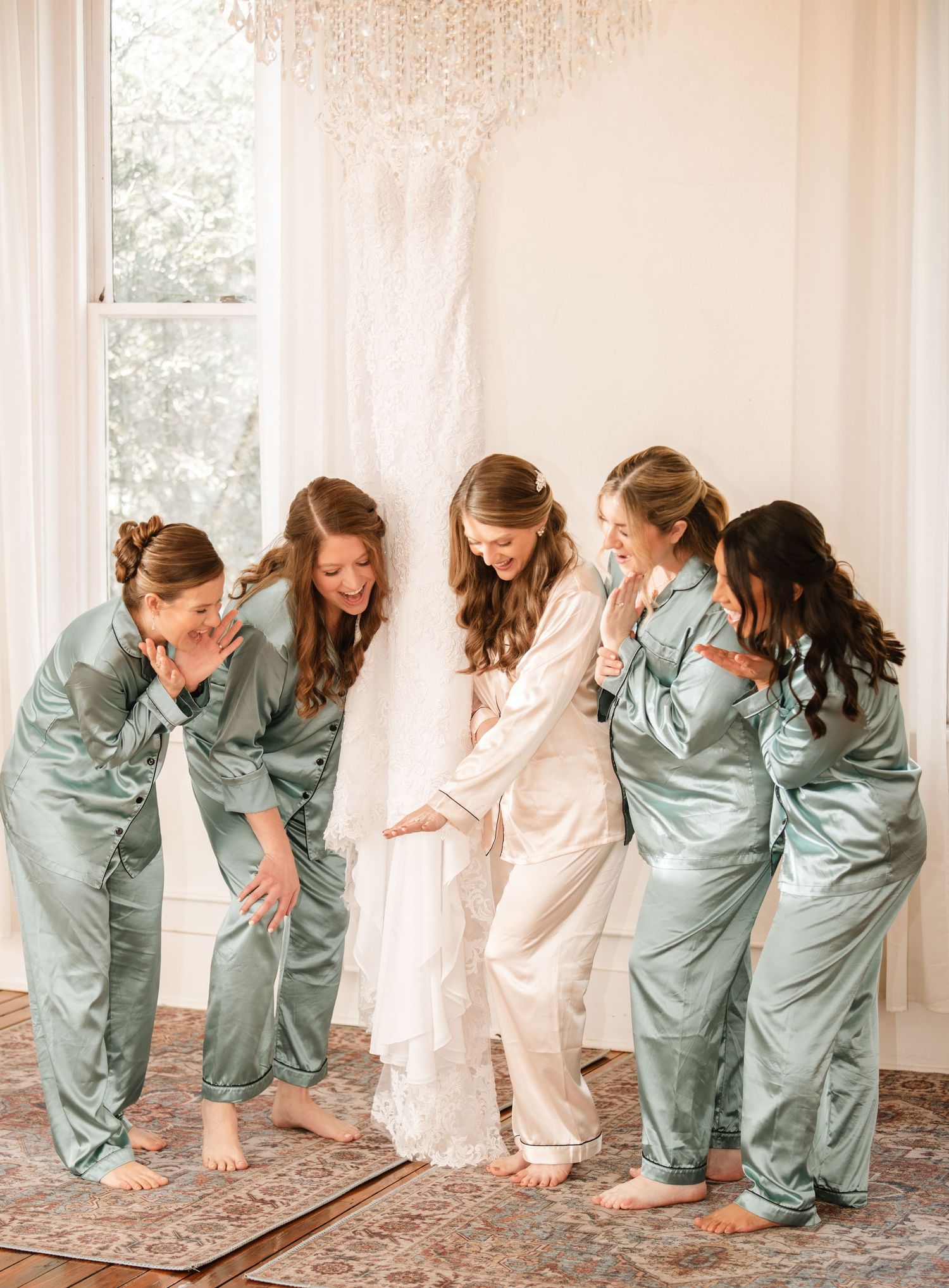 Bridesmaids in sage green satin robes pose playfully while getting ready.