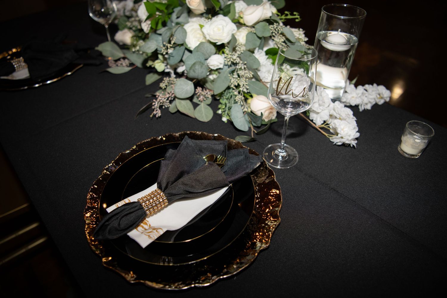 Elegant place settings with gold chargers and white floral centerpieces on black tablecloths.