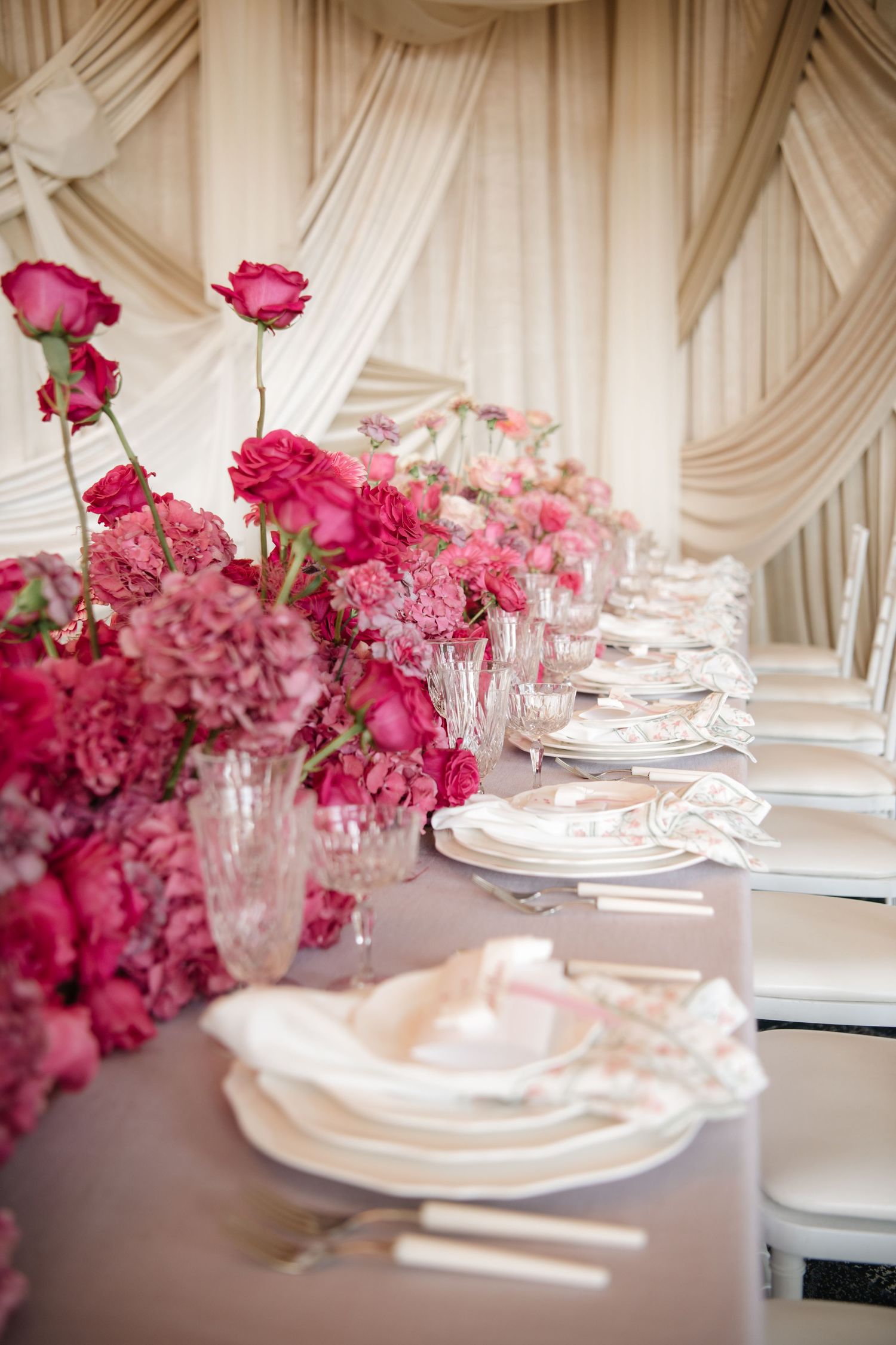 Romantic table setting with pink roses and white place settings against draped fabric background.