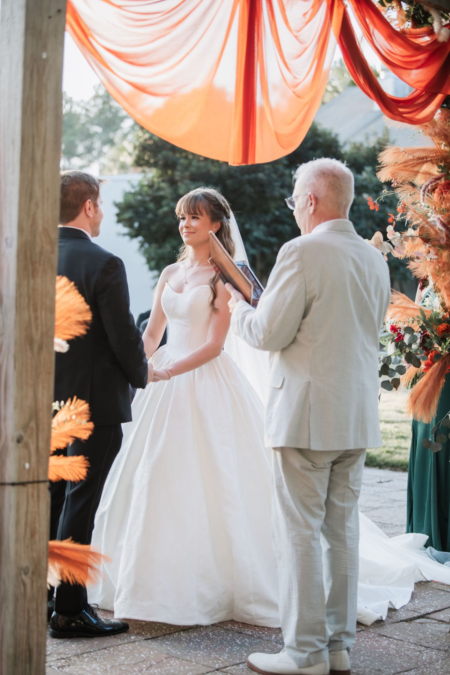 Wedding ceremony under wooden pavilion with coral draping and floral decorations.