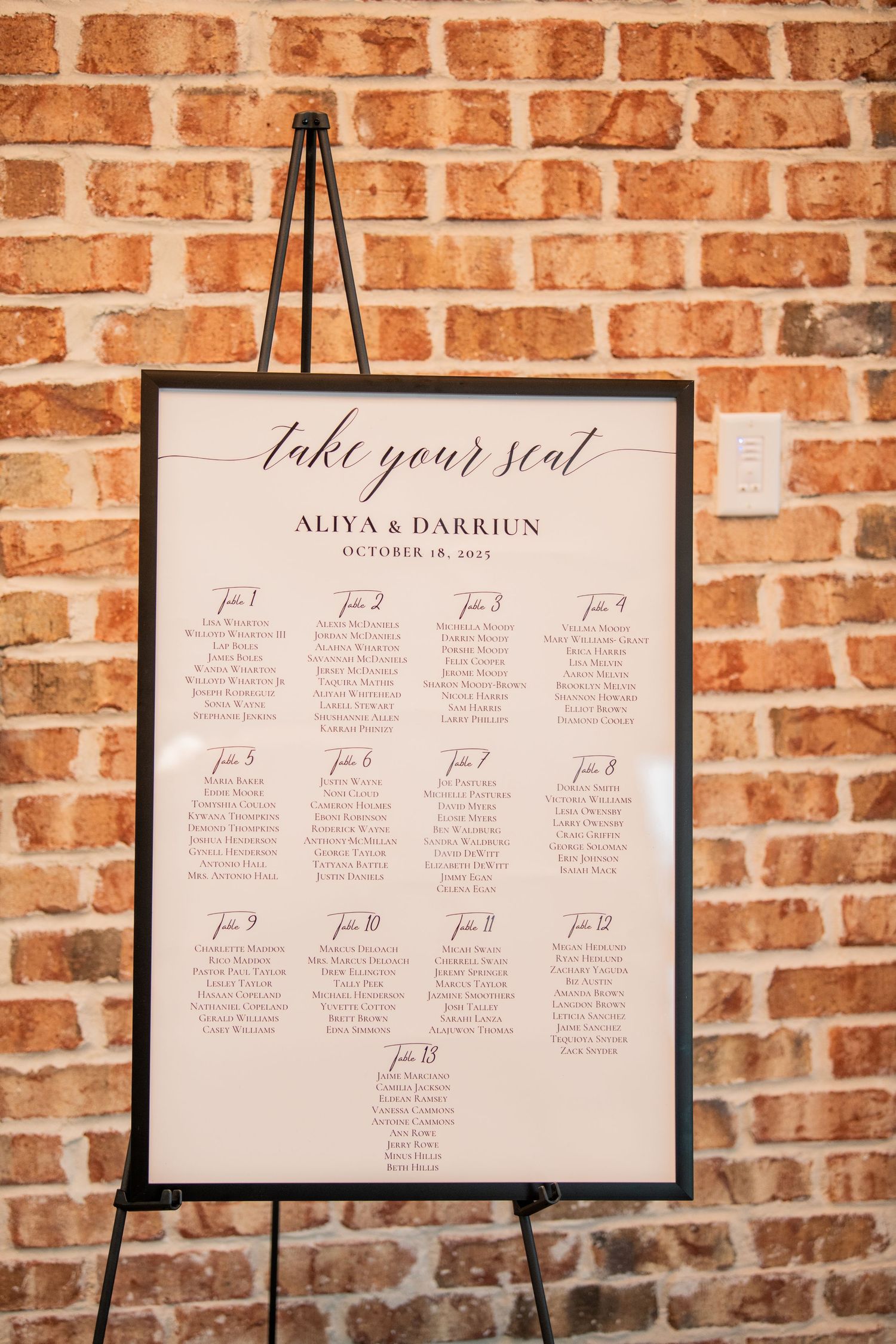 Wedding seating chart displayed on an easel against exposed brick wall.