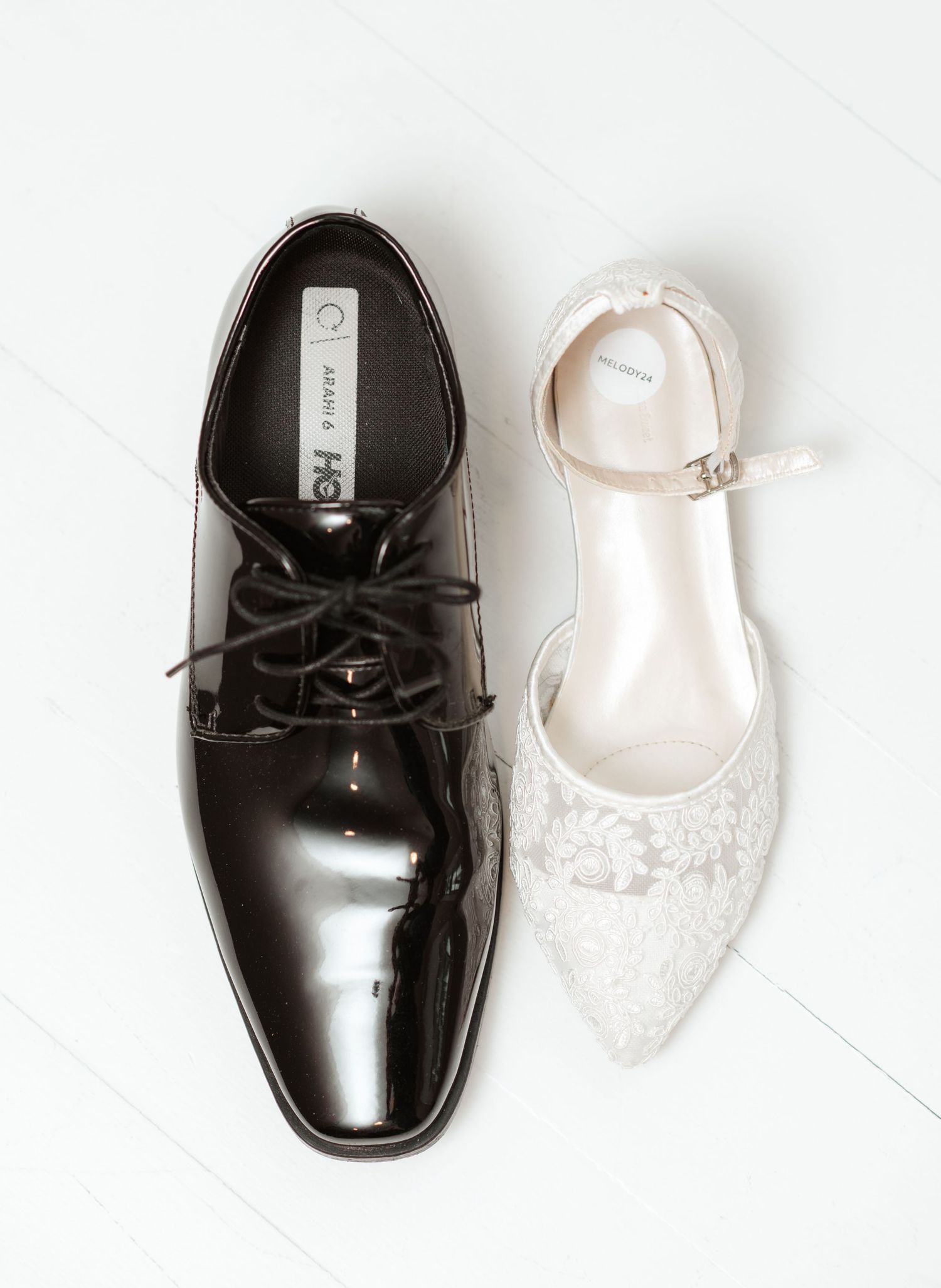 Black patent leather groom's shoe next to white lace bridal shoe on light background.