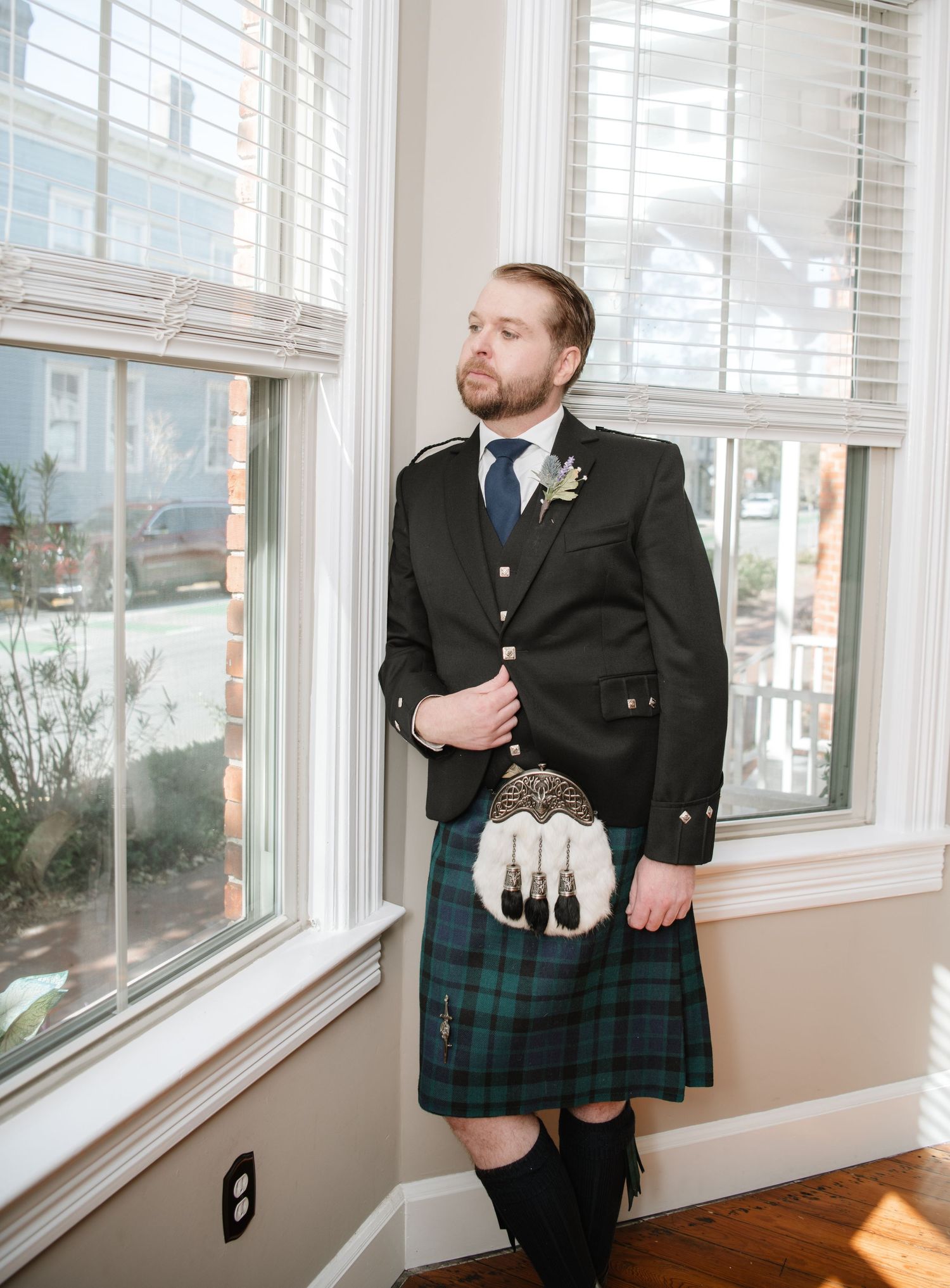 A person wearing a traditional Scottish kilt and formal jacket stands by bright windows.