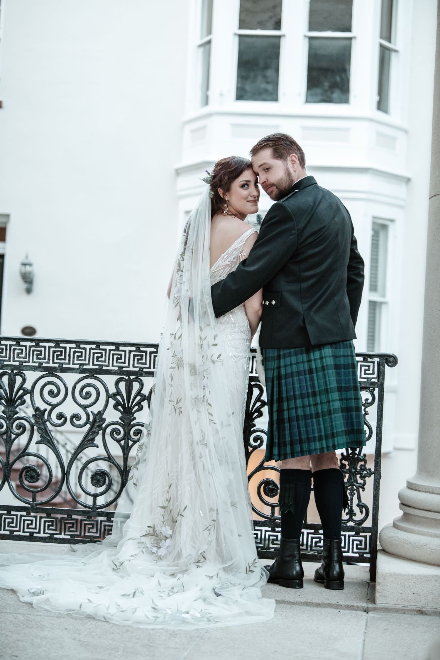 A couple shares an intimate moment on a white balcony with decorative iron railings.