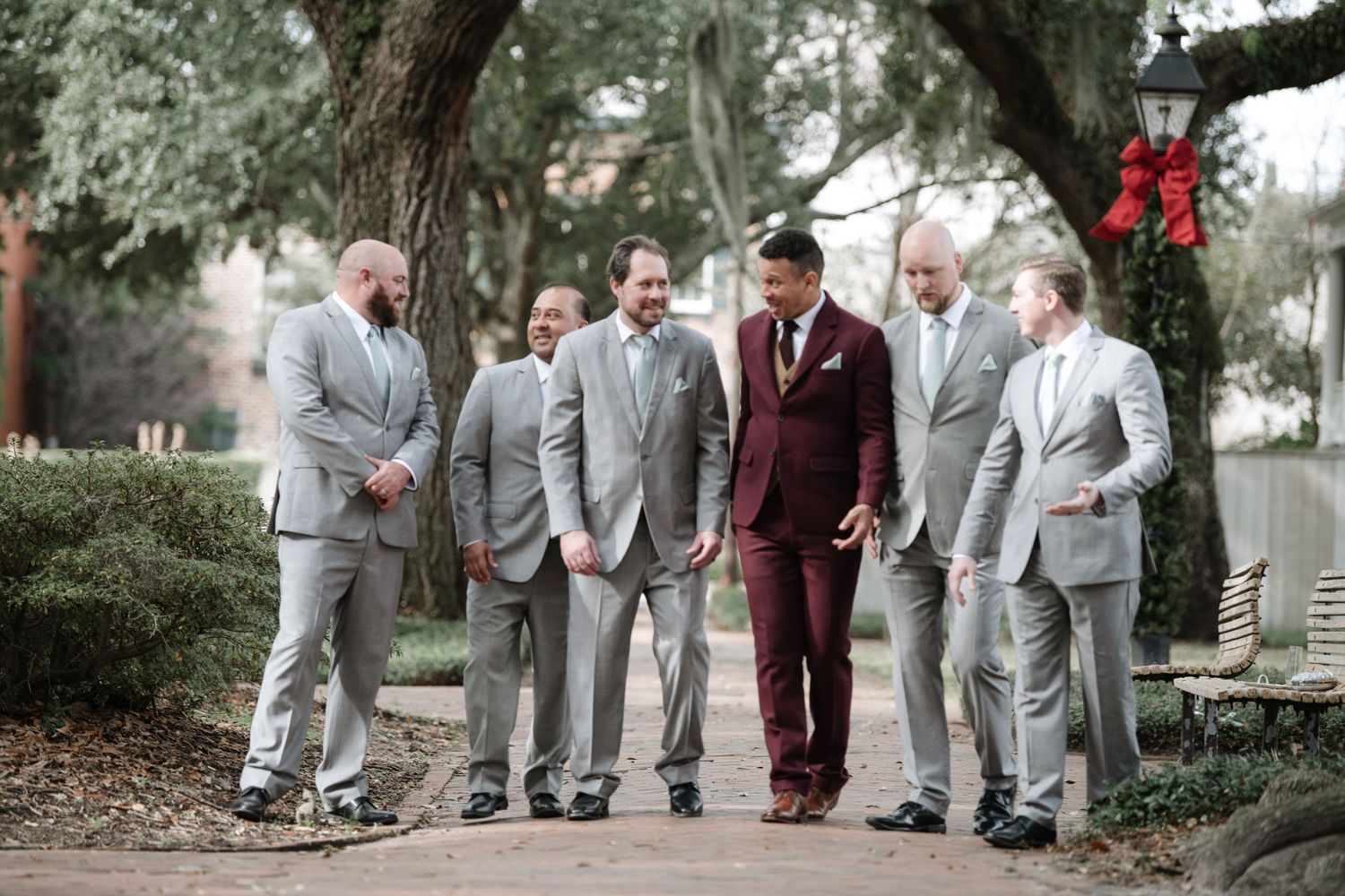 Groomsmen in light gray suits and one in burgundy stand together in an outdoor garden setting.