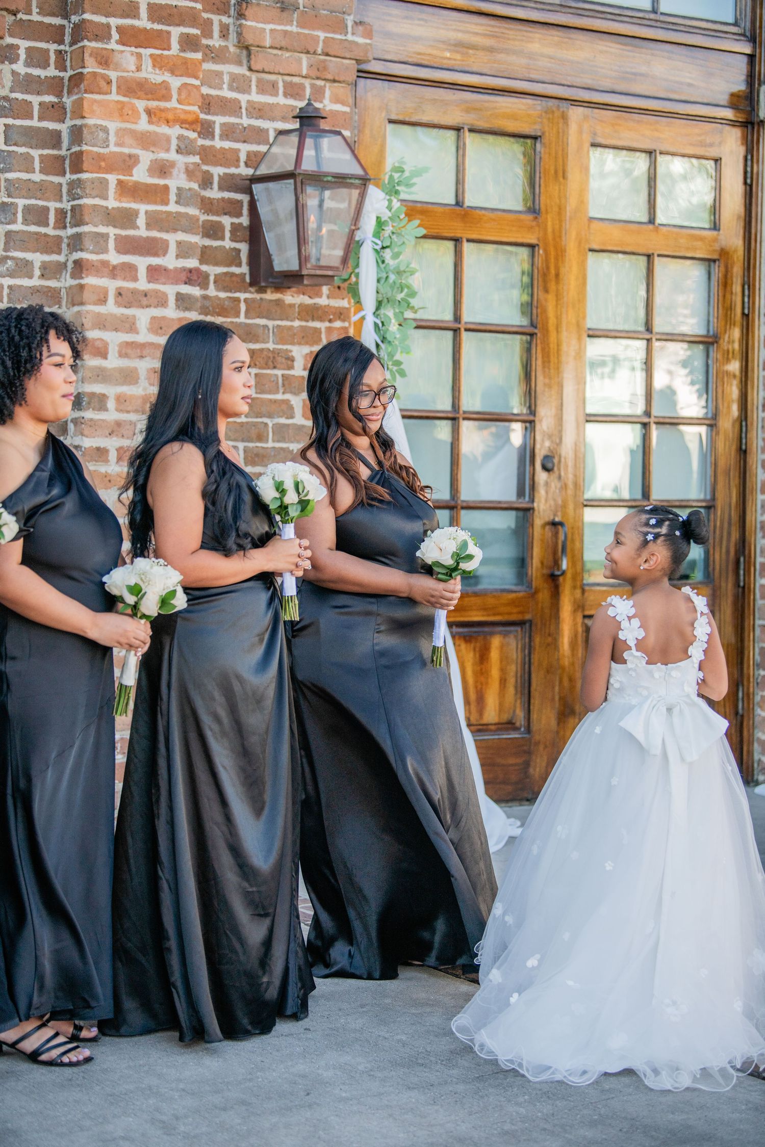 Bridesmaids in elegant black dresses stand with a young flower girl in a white gown by a rustic brick entrance.