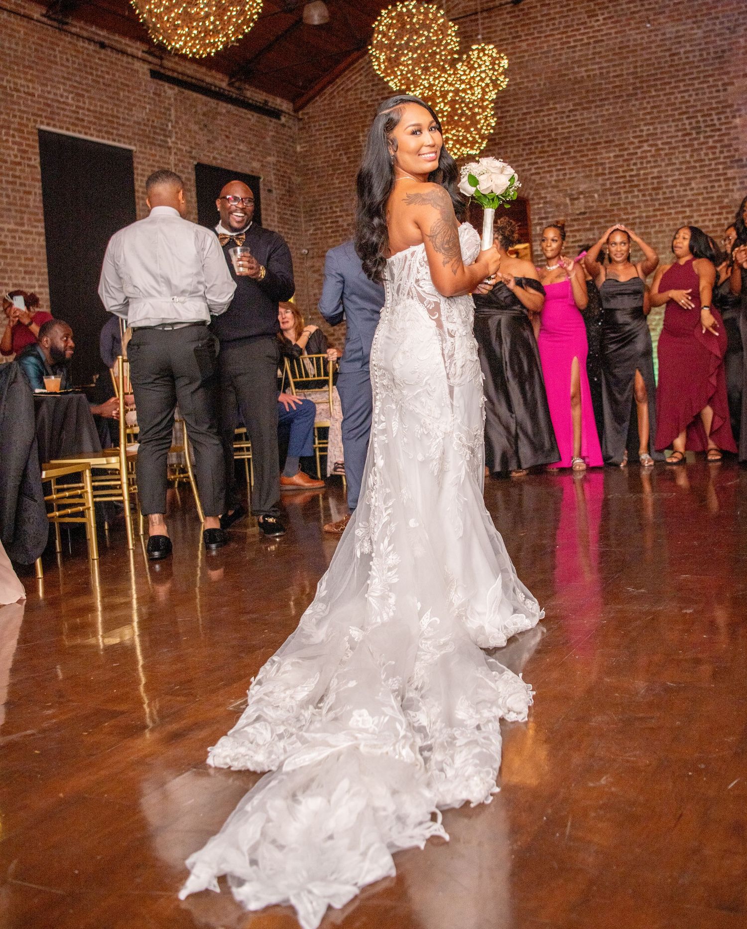 Bride in flowing white lace dress dances at wedding reception with gold chandeliers and brick walls.