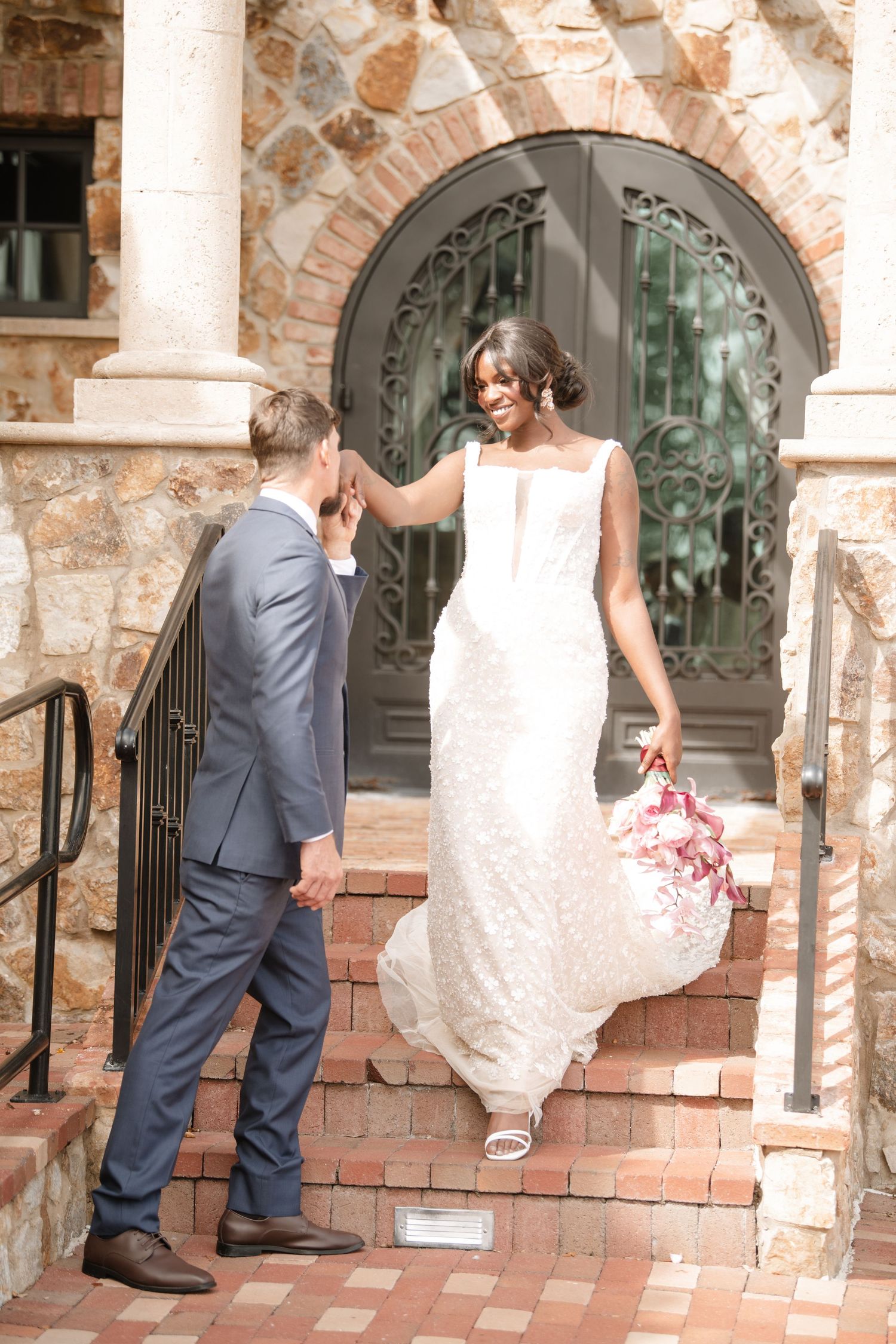 Couple in wedding attire interacts on brick steps near stone columns and arched windows.
