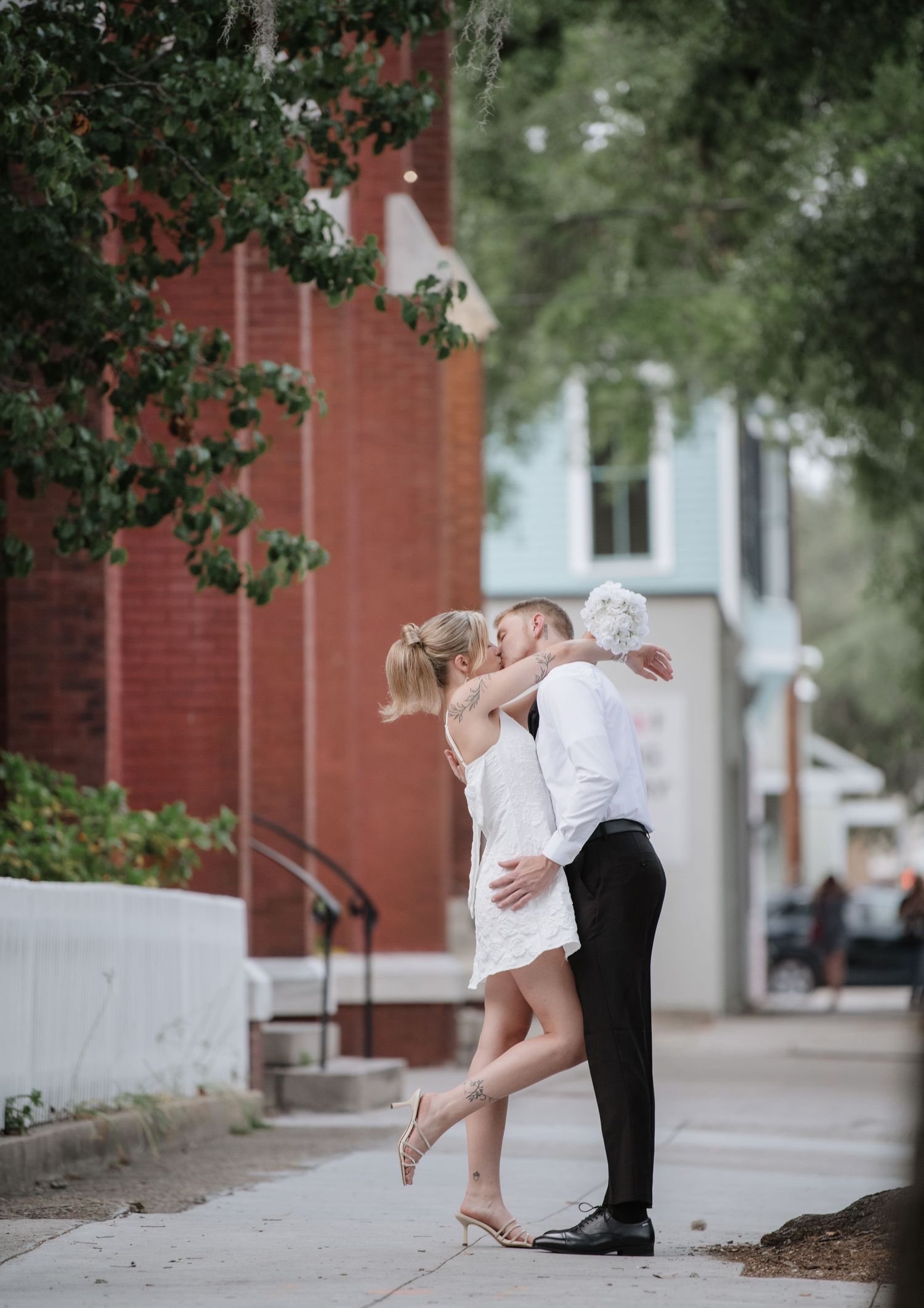 A romantic moment between a couple on a city sidewalk with overhanging trees.