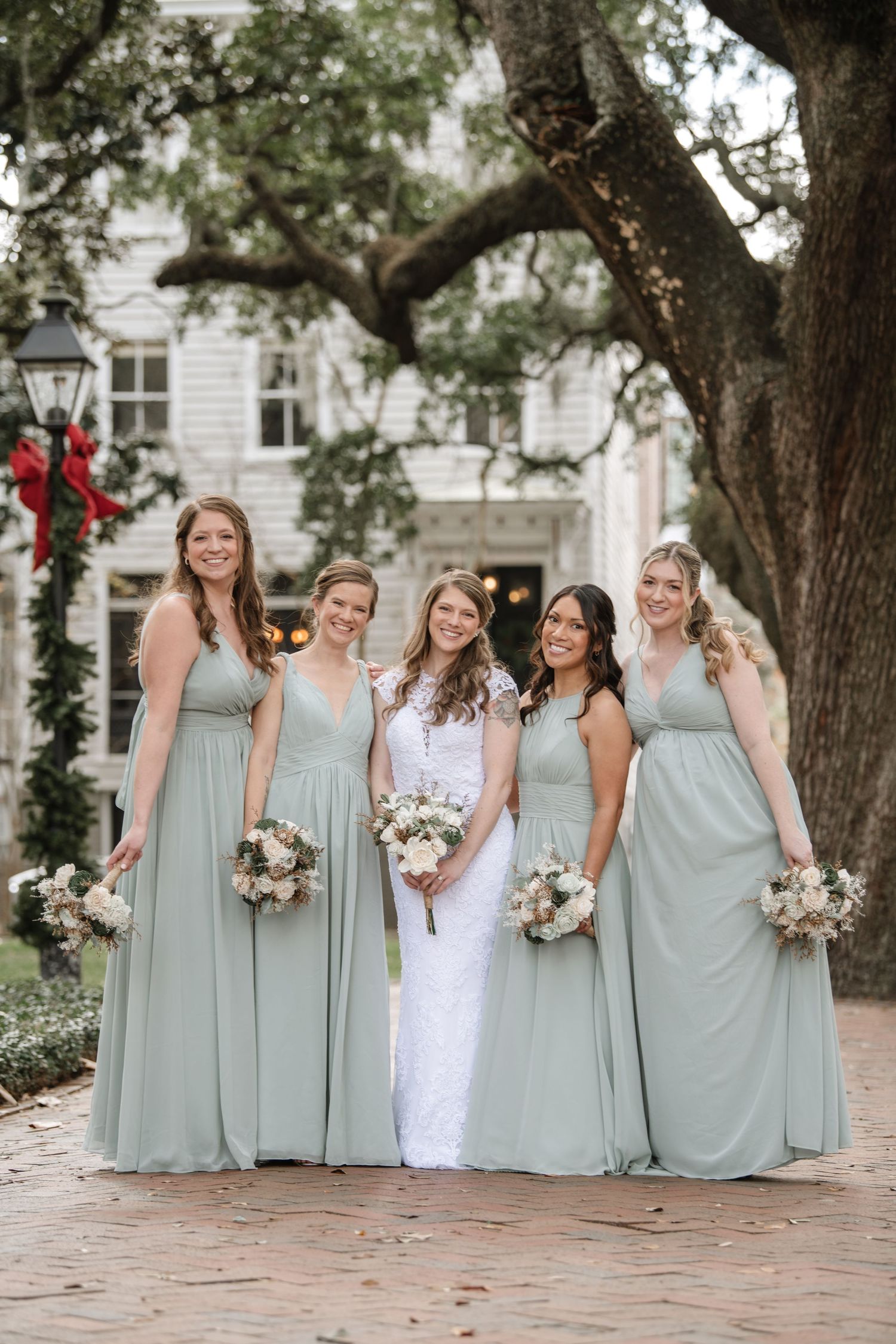 Bridesmaids in sage green dresses pose together outdoors while holding bouquets.