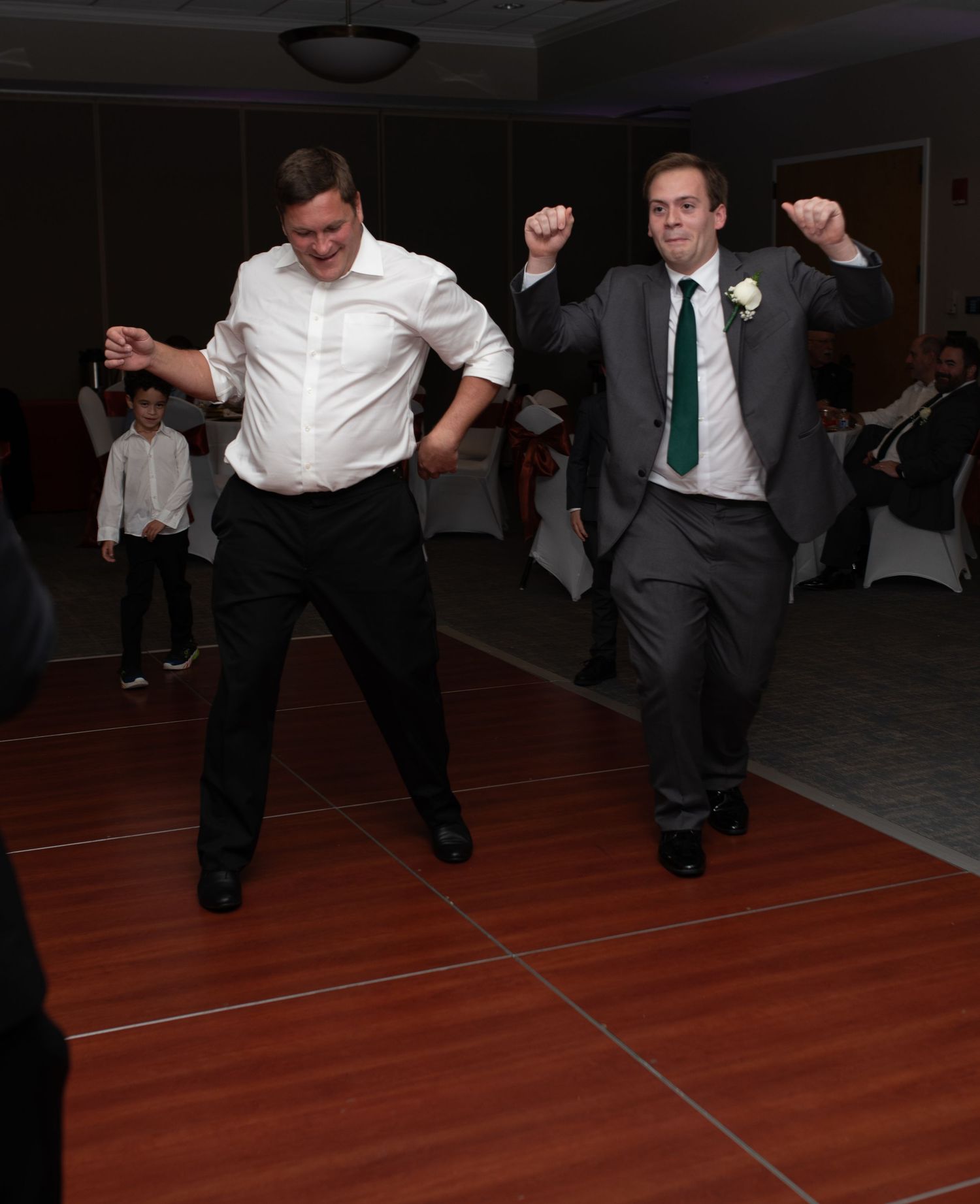 Two men dancing energetically on a wooden dance floor at a wedding reception.