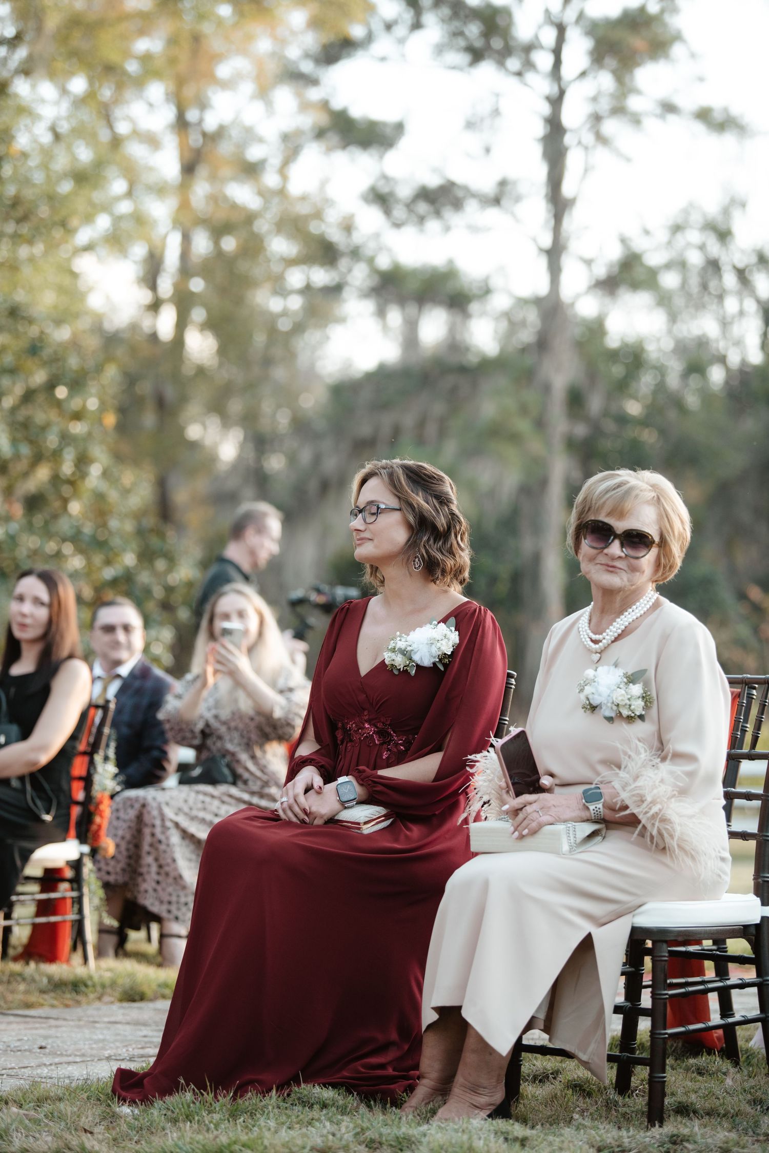 Wedding guests in burgundy and cream colored dresses seated on wooden chairs at an outdoor ceremony.
