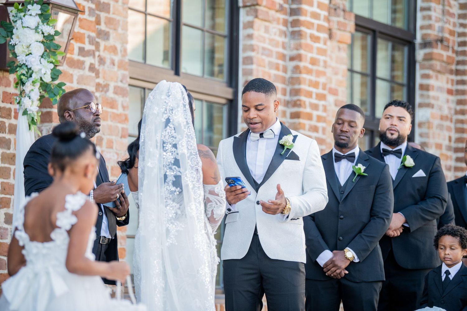 Wedding party members pose in formal attire outside historic brick building.