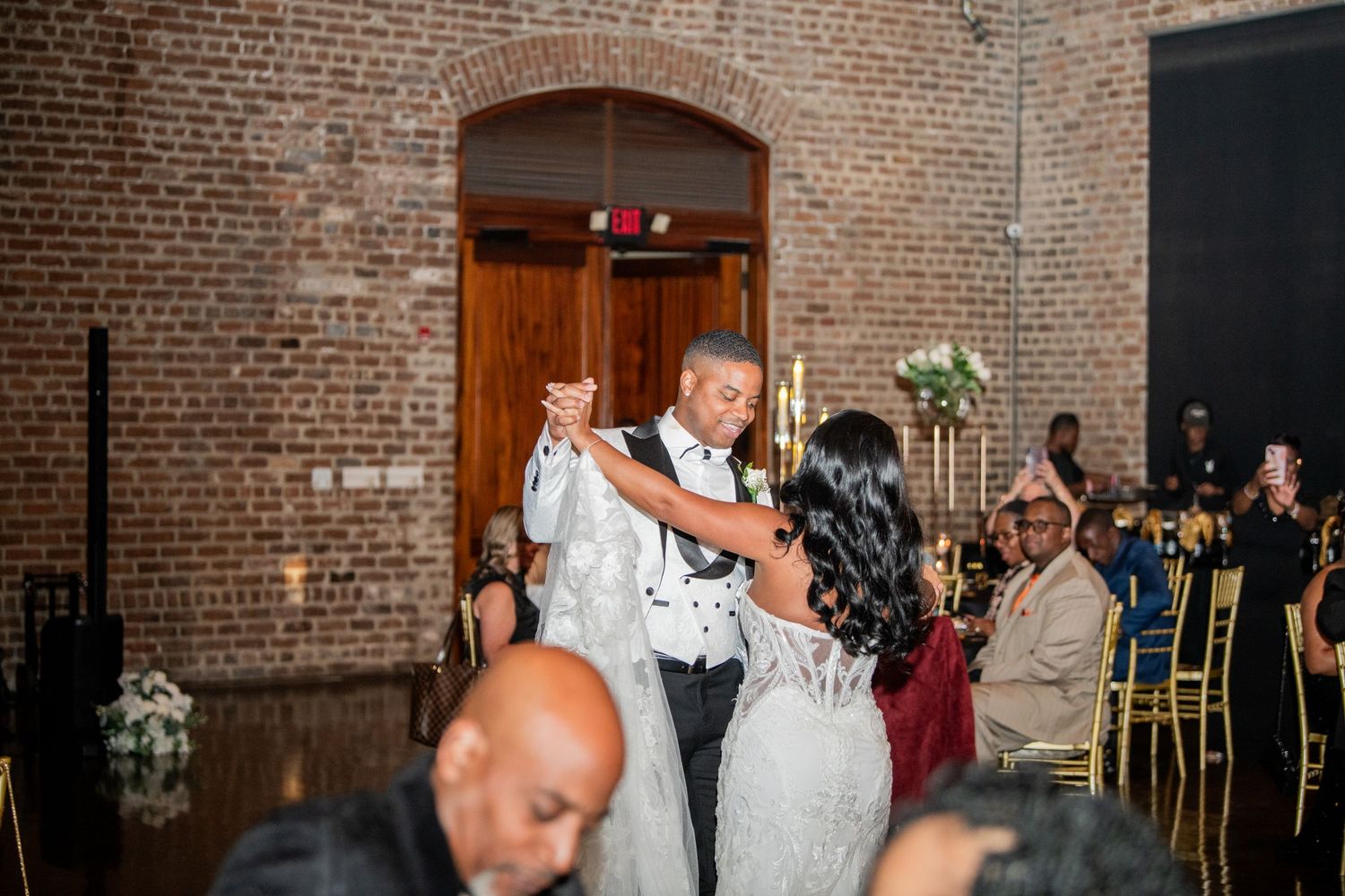 Couple sharing their first dance at a rustic brick-walled wedding venue with guests watching.