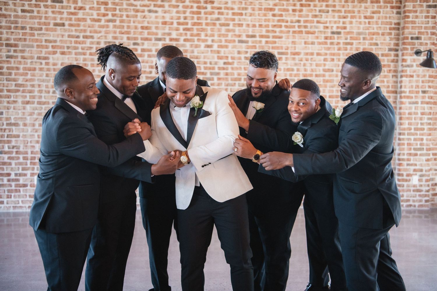 Groomsmen share joyful moments and laughter in formal attire against a brick wall backdrop.