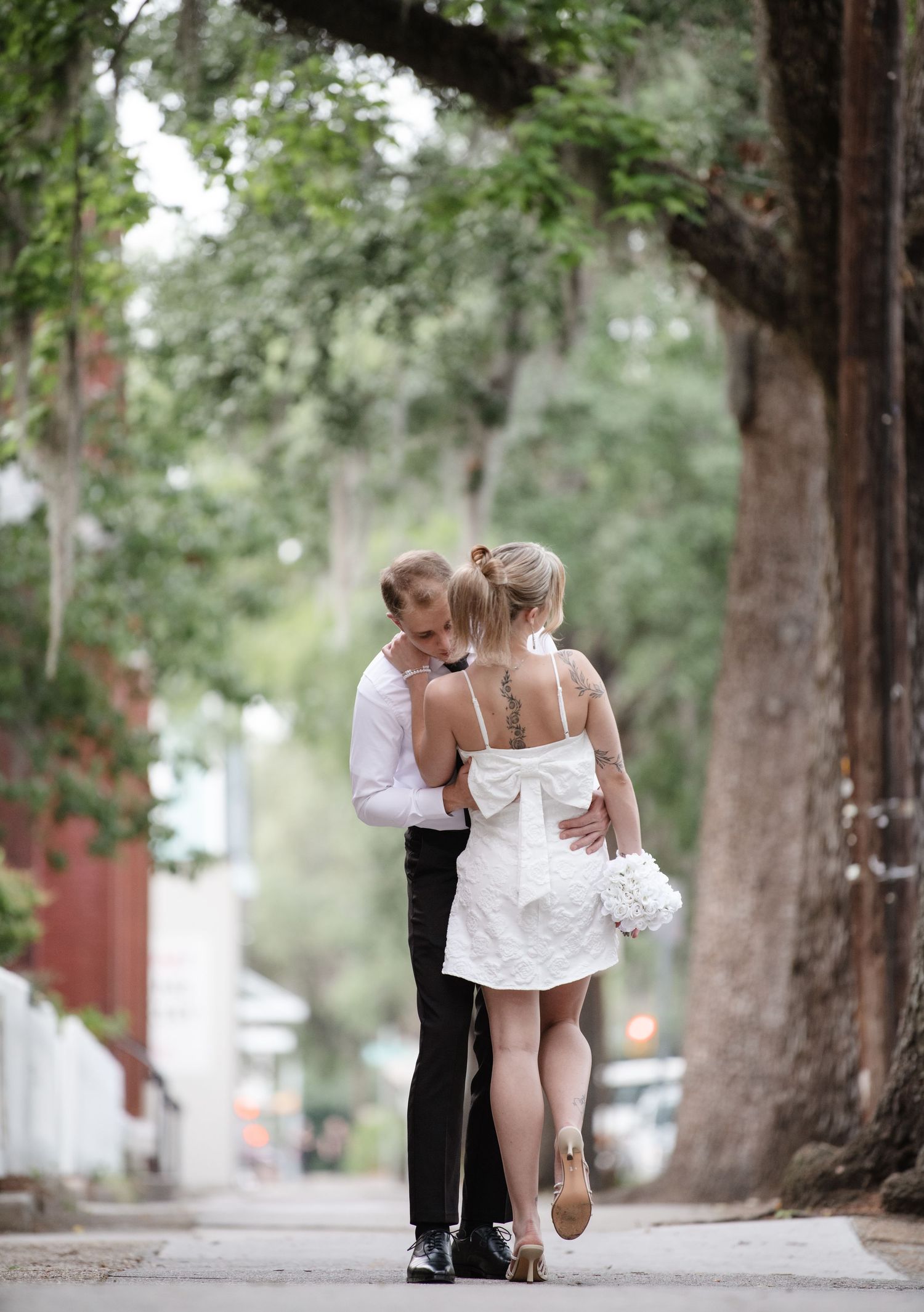 A romantic couple walking together on a tree-lined sidewalk with brick buildings in the background.